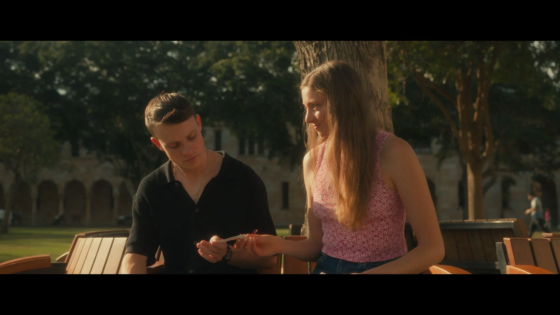Two teenagers, a boy and a girl, sitting on a park bench under a tree, looking at a smartphone together.