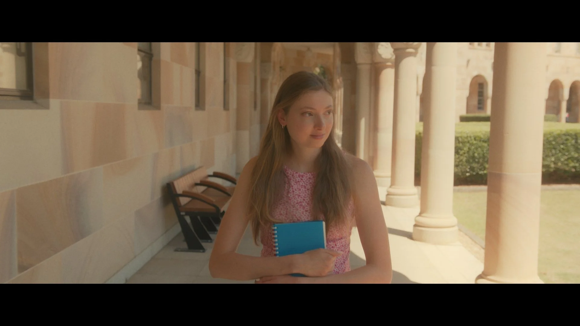 A young woman with long, wavy hair standing in a covered outdoor walkway, holding a blue notebook, looking to her right, with a background of large stone columns and a manicured hedge.