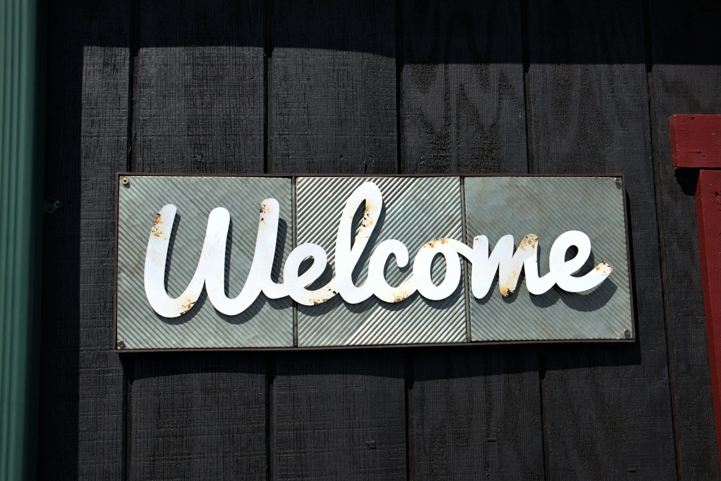A close-up photograph of a sign with the word 'welcome' written in white cursive letters, mounted on a black wooden wall with visible texture and grain.