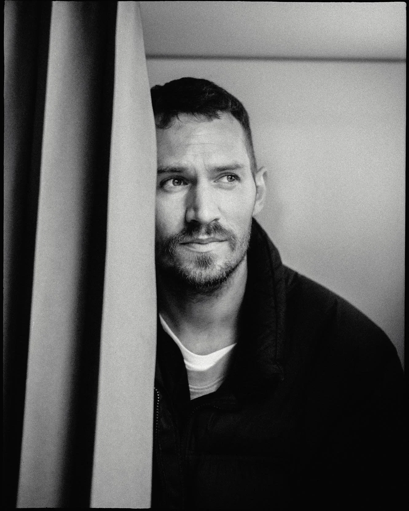A black-and-white photo of a man with short dark hair, a beard, and mustache, looking to the side with a thoughtful expression. Part of his face is obscured by a curtain or fabric on the left side of the image.