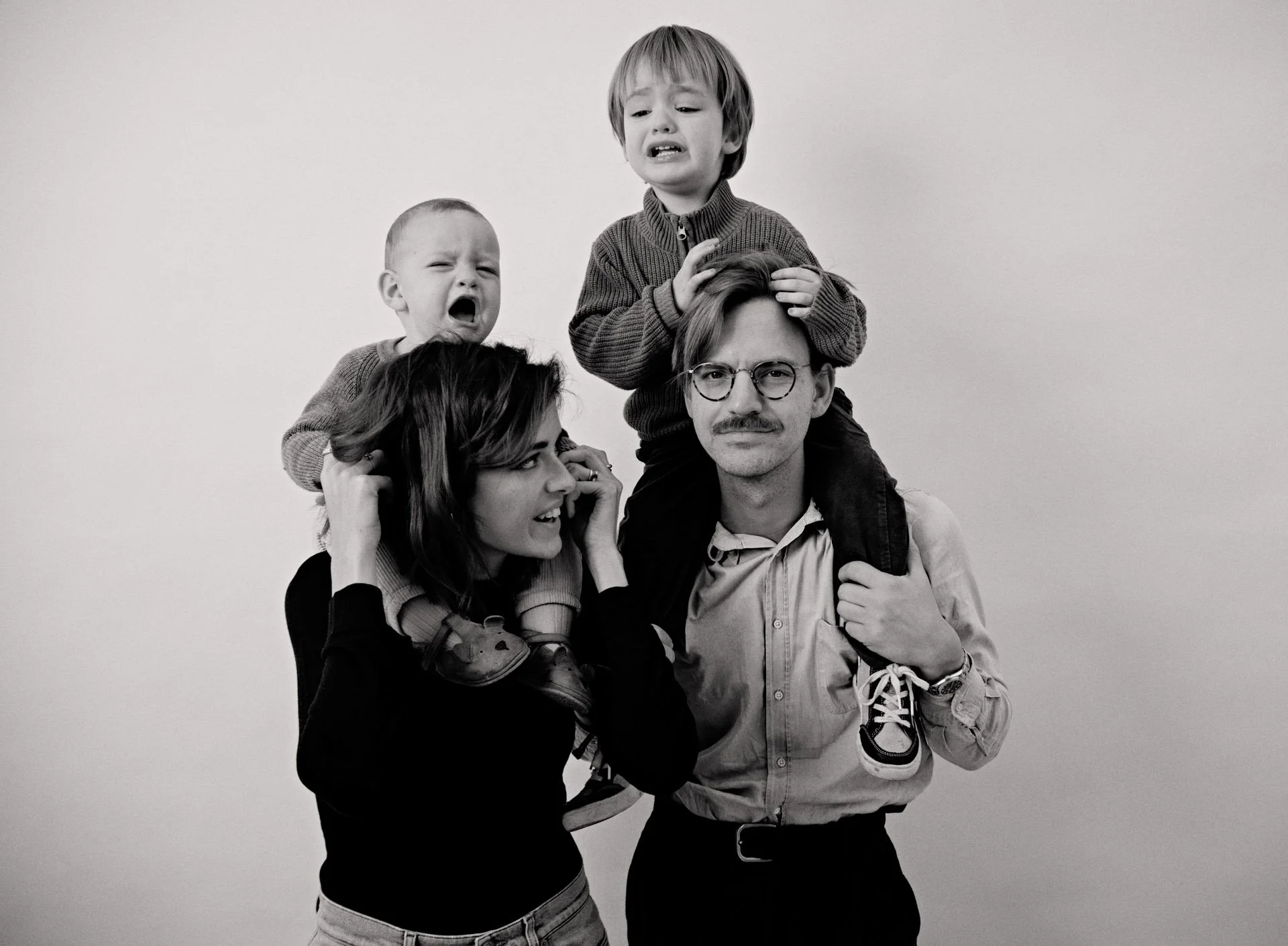 Black and white photo of a man with glasses, a woman, and two children. The man is carrying one child on his shoulders and holding the other child's foot. The woman is holding onto the child's foot and smiling, while the children have distressed and 