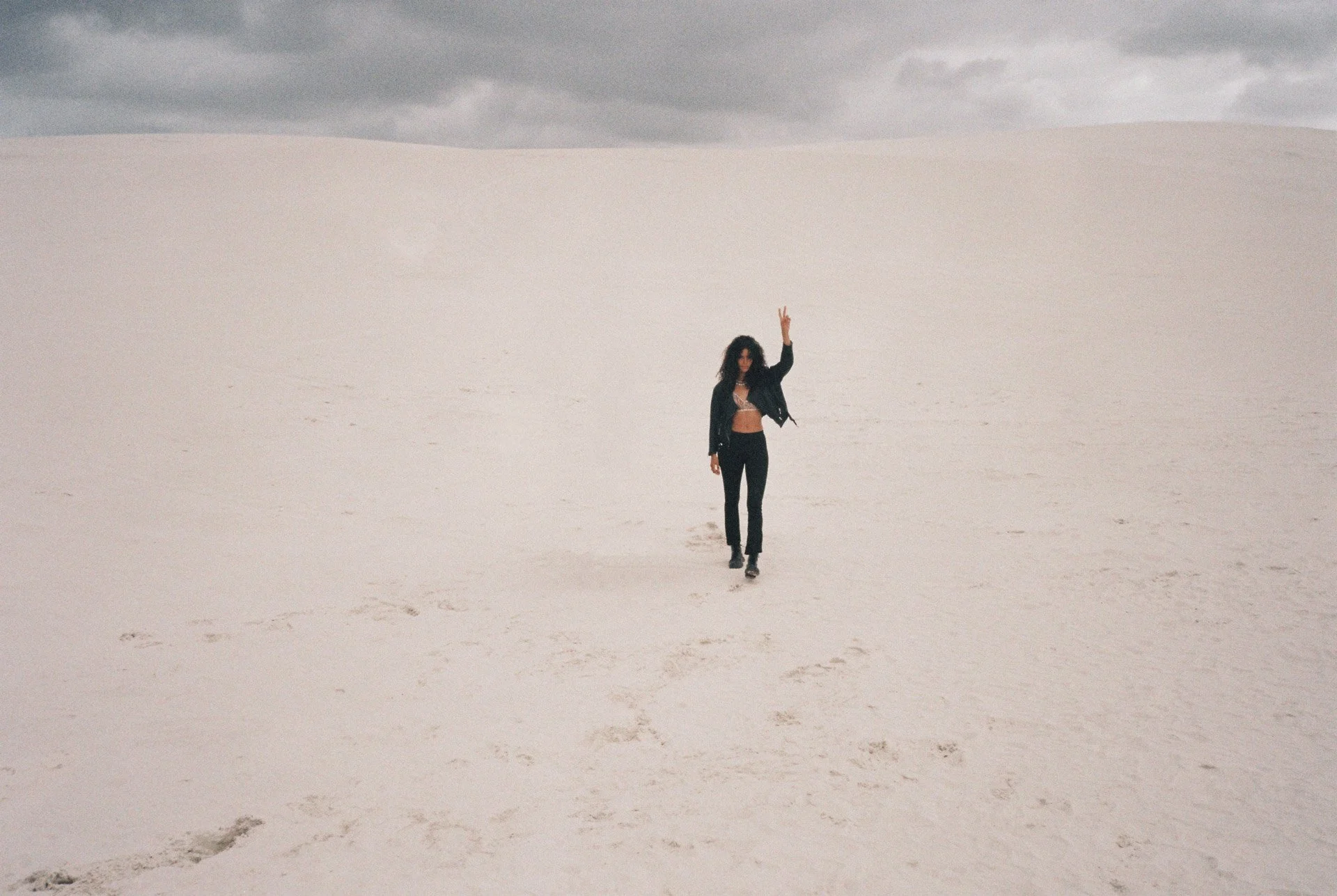 A woman standing in a vast, white desert with cloudy skies, making a peace sign with her right hand.