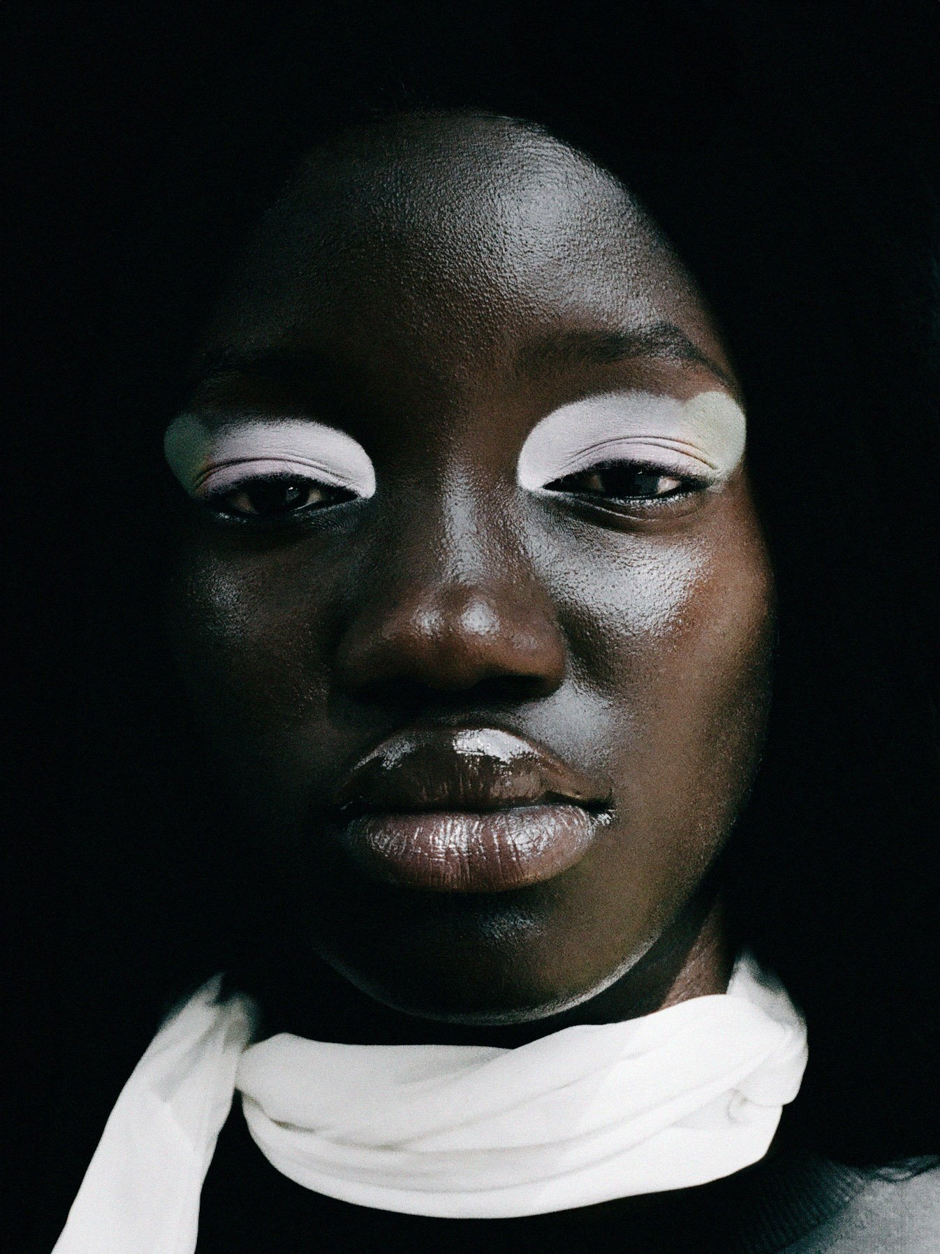 Close-up of a person with dark skin, white eye makeup, and glossy lips, wearing a white cloth around their neck.