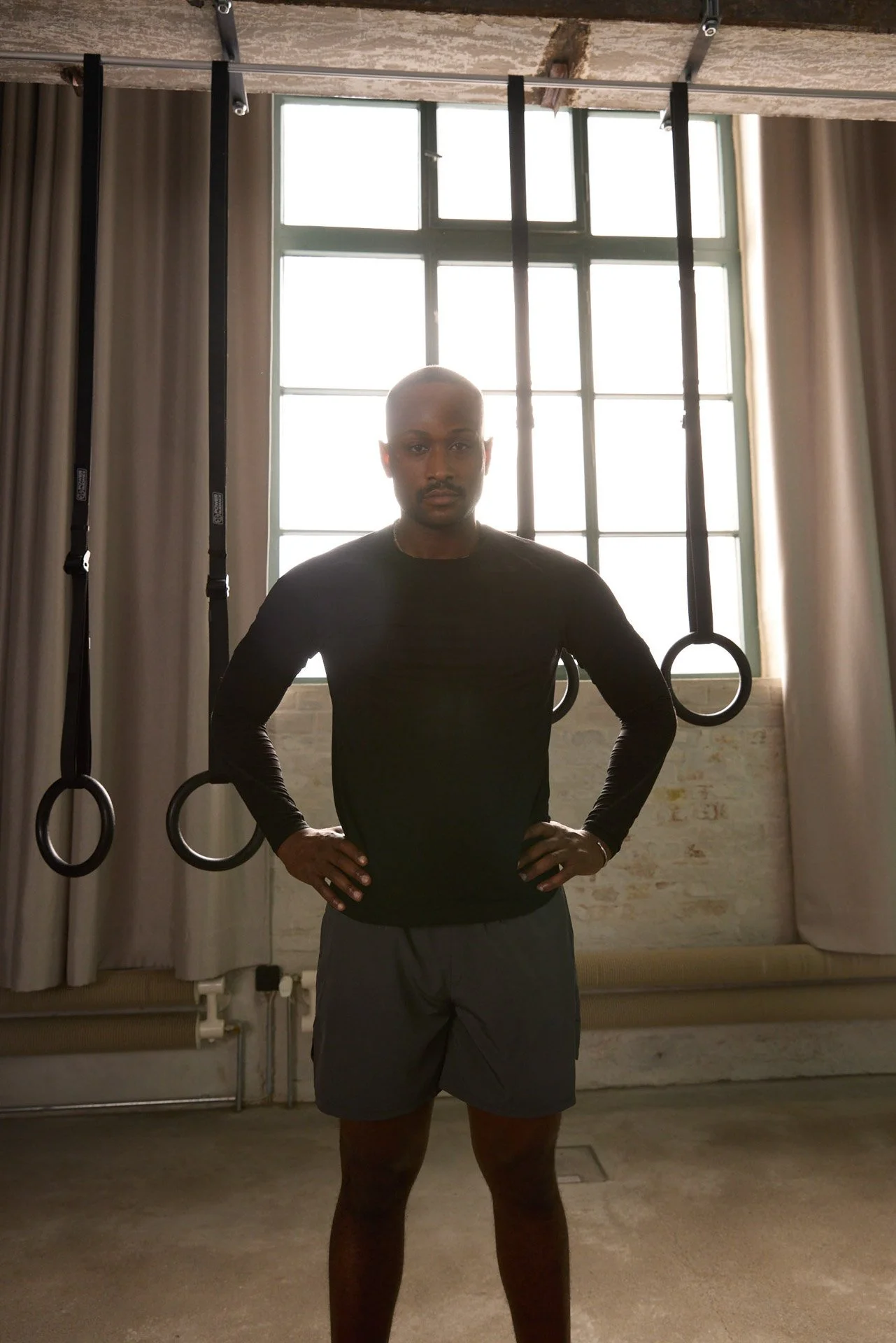 A man standing with hands on hips in front of a large window with gymnastic rings hanging from the ceiling.