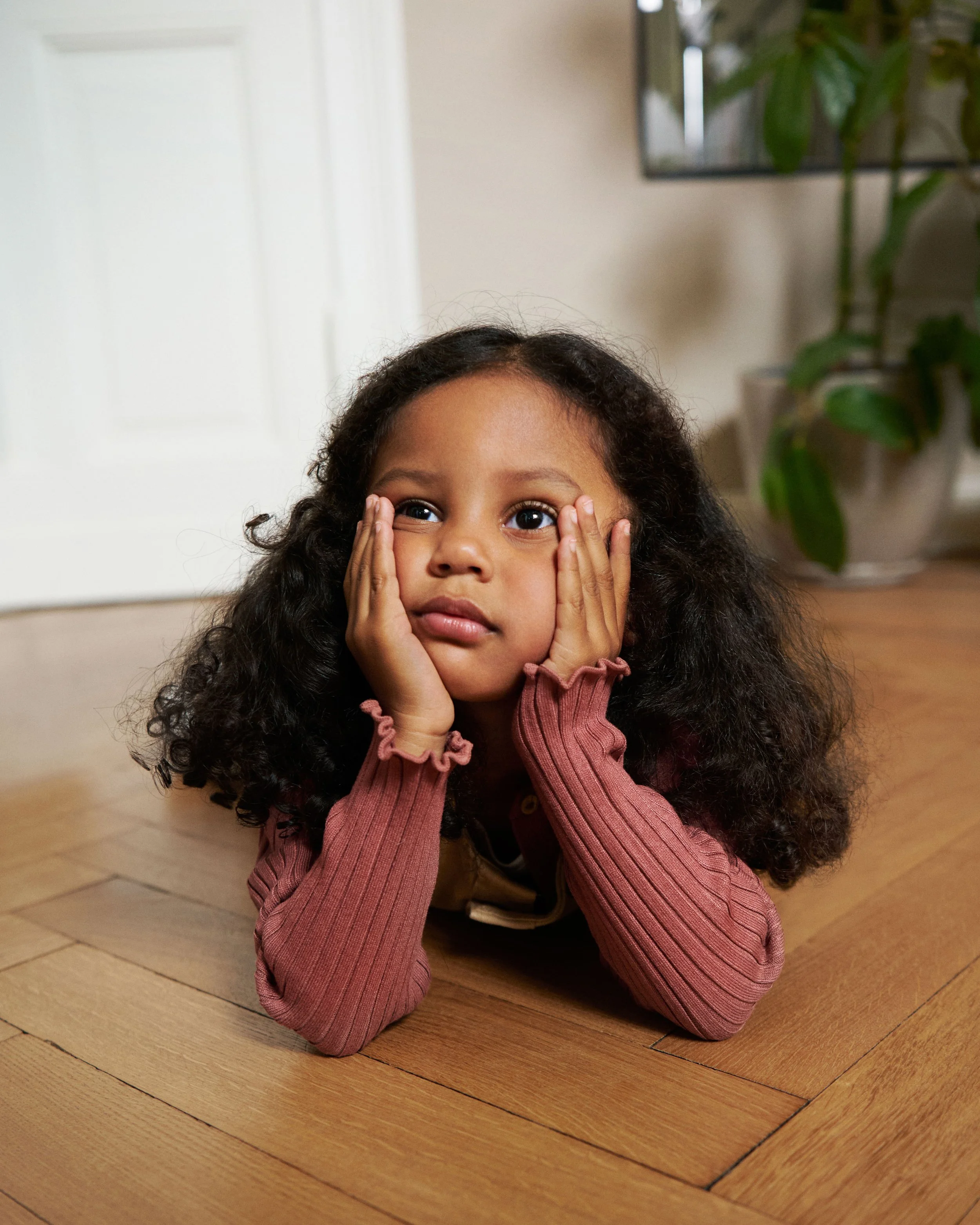 A young girl with curly hair and brown skin lies on a wooden floor, resting her face in her hands, gazing thoughtfully.