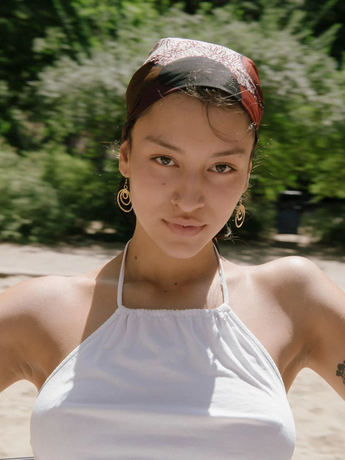 A young woman with short hair wearing earrings, a headscarf, and a white halter top, standing outdoors with a blurred green background.