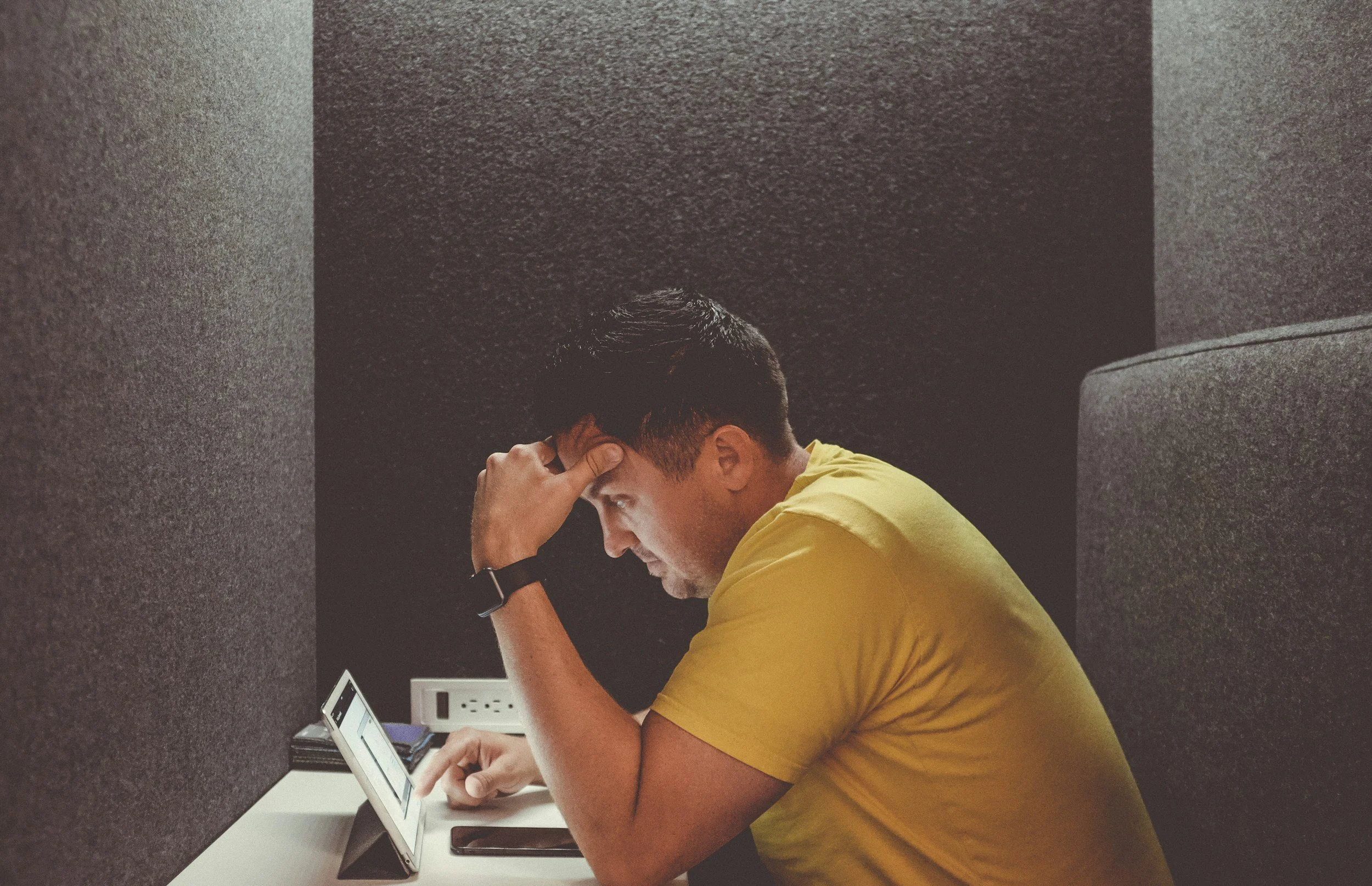 A young man wearing a yellow shirt sitting at a desk with a tablet. He appears to be stressed or deep in thought, resting his head on his hand. The desk is in a confined, dark gray space with a power outlet and some electronic devices.