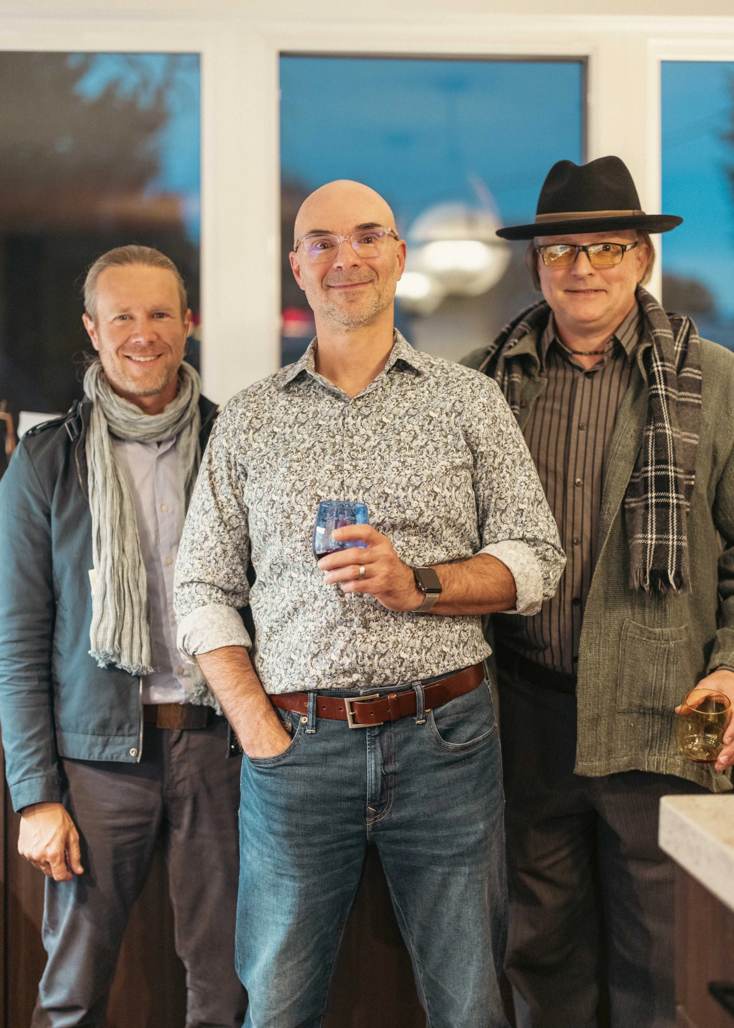 Three men standing together at a gathering, smiling for the camera. One man in the center holds a glass of red wine.