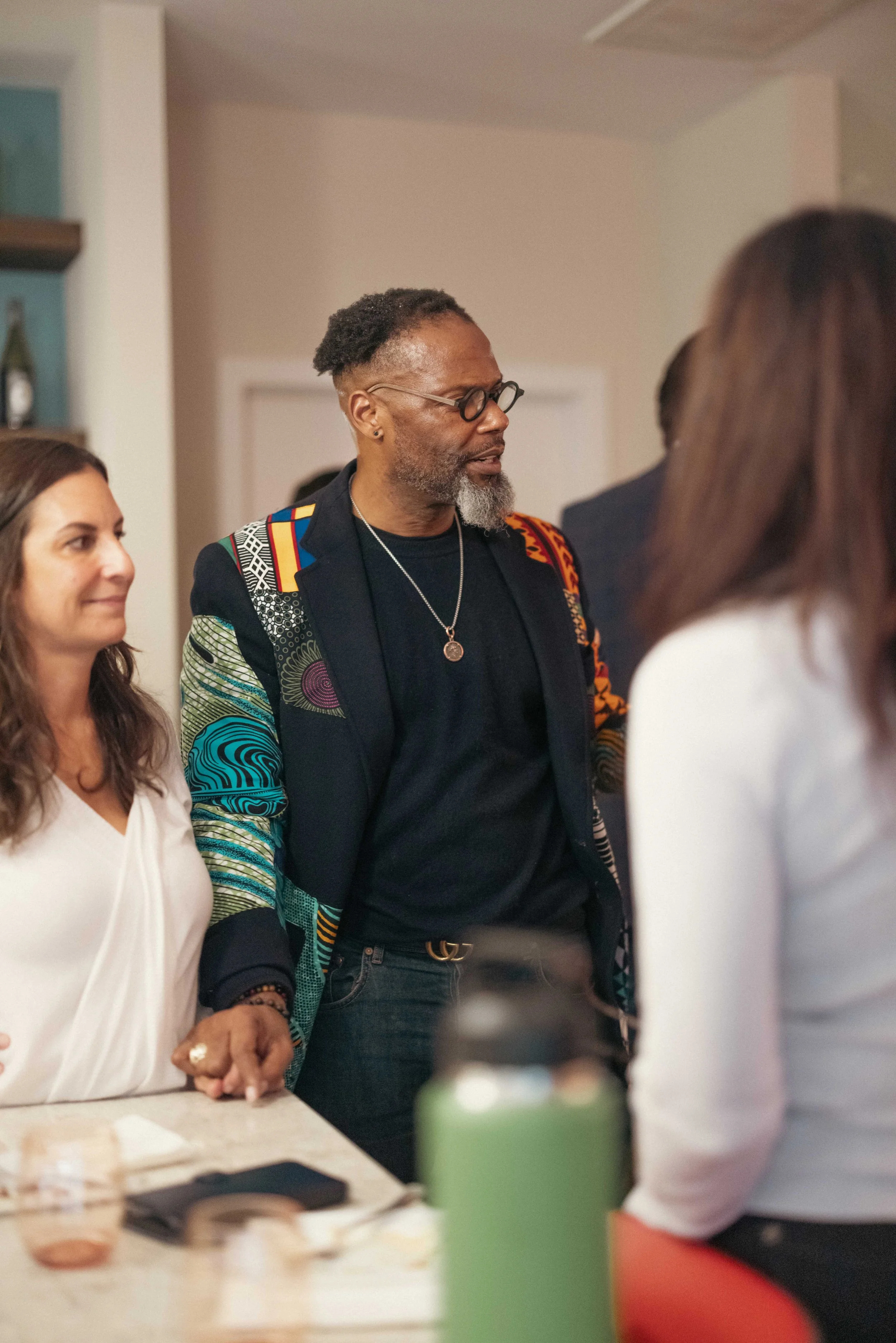 A group of diverse people gathered indoors, engaging in conversation. The image features a woman with long brown hair, a man with glasses and gray beard dressed in a colorful patterned jacket, and other individuals in the background.