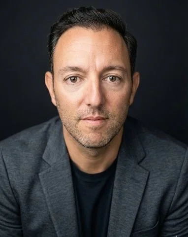 Headshot of a man with dark hair, wearing a dark blazer and black shirt, against a dark background.