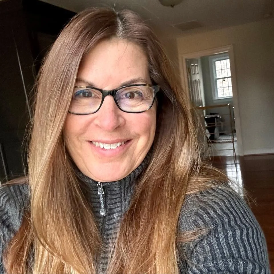 A woman with long, straight light brown hair and glasses smiling in a room with wooden floors, a window, and a doorway in the background.