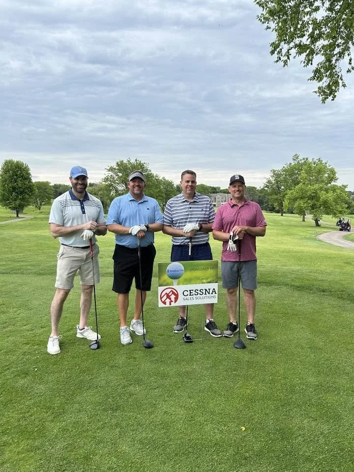 Four men standing on a golf course holding golf clubs, with a sign for Cessna Sales Solutions in front of them, under a cloudy sky.