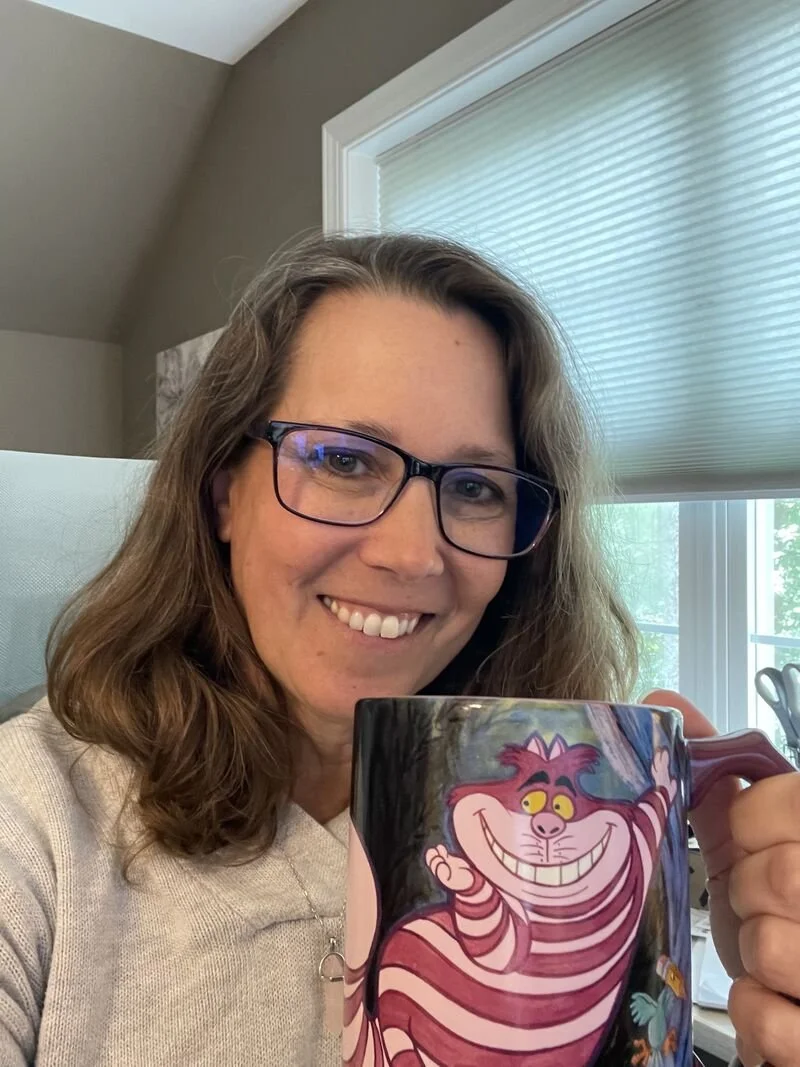 A woman smiling while holding a colorful mug with a cartoon Cheshire Cat illustration from Alice in Wonderland.