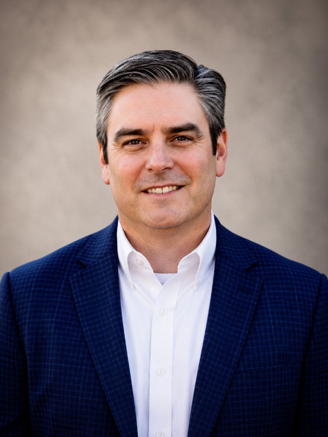 A headshot of a man with dark hair styled to the side, wearing a dark blue checkered blazer over a white collared shirt, smiling slightly, against a neutral background.