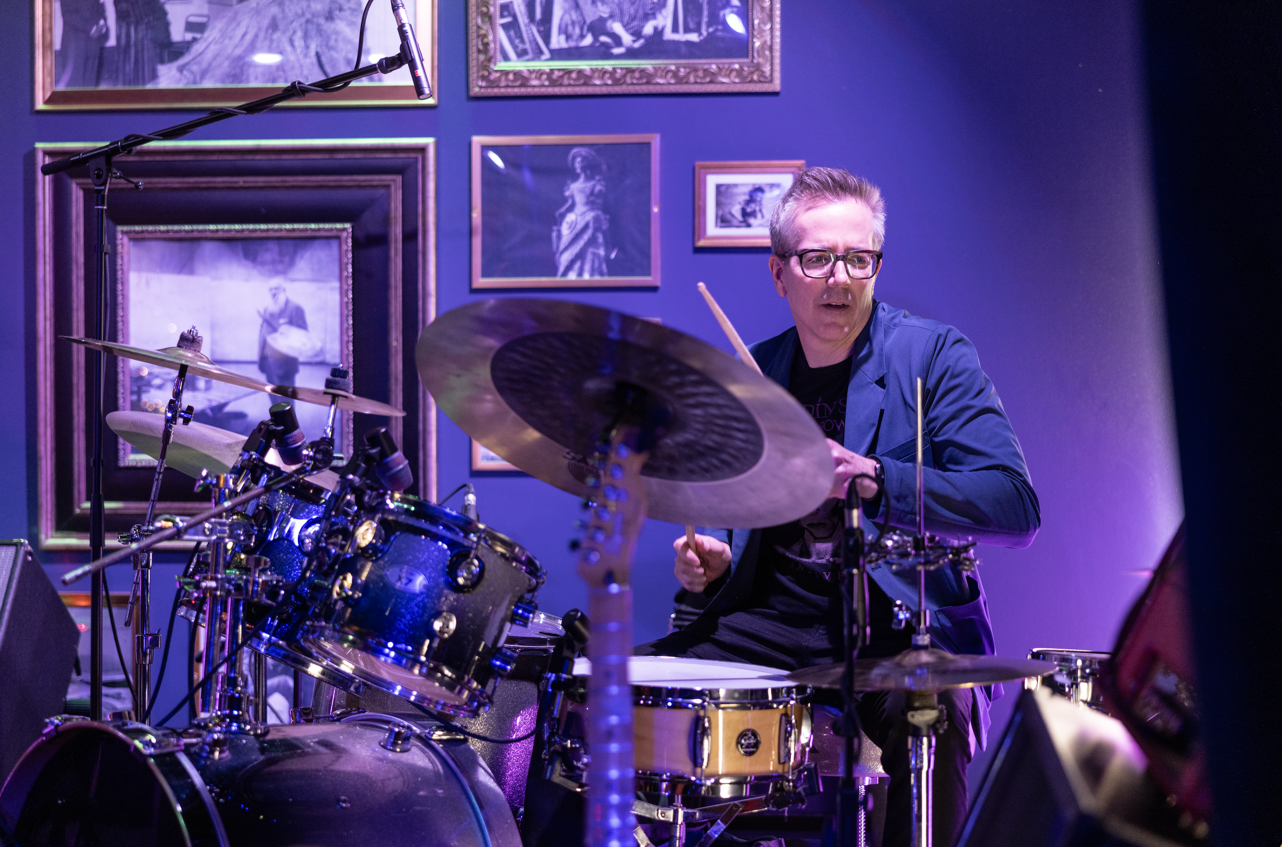 A man with gray hair and glasses playing drums during a live performance in a dimly lit room with purple lighting, decorated with framed artwork on the wall.