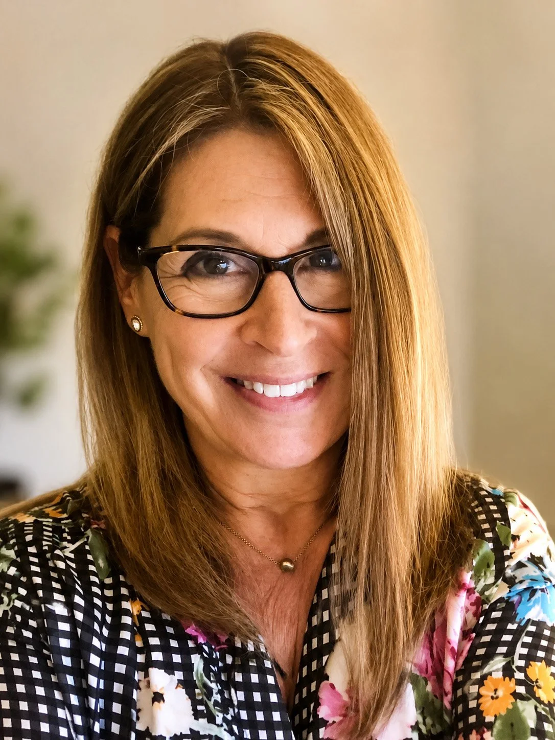 A woman with shoulder-length light brown hair, glasses, and earrings, smiling and wearing a floral and checkered shirt.