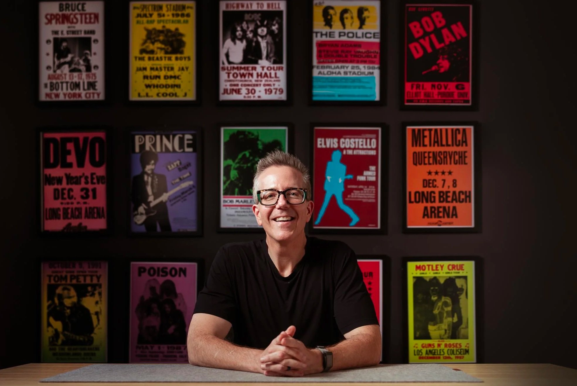 A man with glasses and a black shirt is sitting at a wooden table, smiling, with his hands clasped. Behind him is a dark wall adorned with framed posters of various music artists and concerts, including Bruce Springsteen, The Beastie Boys, The Police, Bob Dylan, Elvis Costello, Metallica, and others, in vibrant colors.