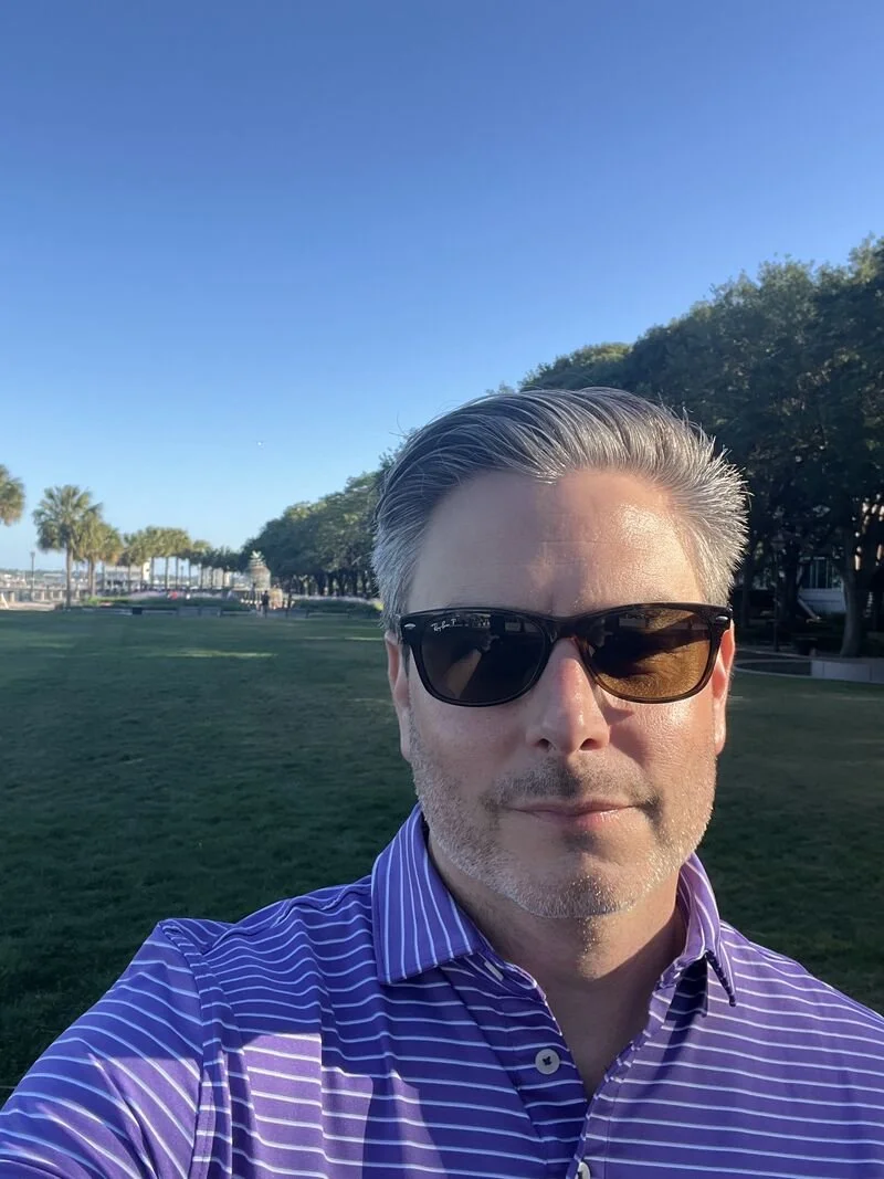 A man wearing sunglasses and a purple striped dress shirt taking a selfie outdoors on a sunny day, with green grass, trees, and a clear blue sky in the background.