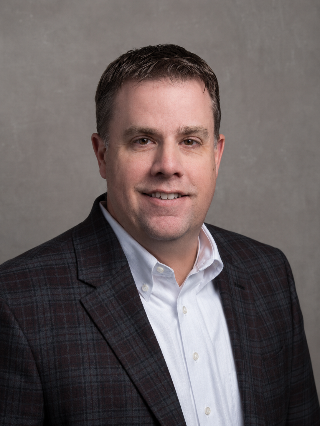 Professional headshot of a man with short brown hair, wearing a dark plaid blazer over a white shirt, against a plain gray background.