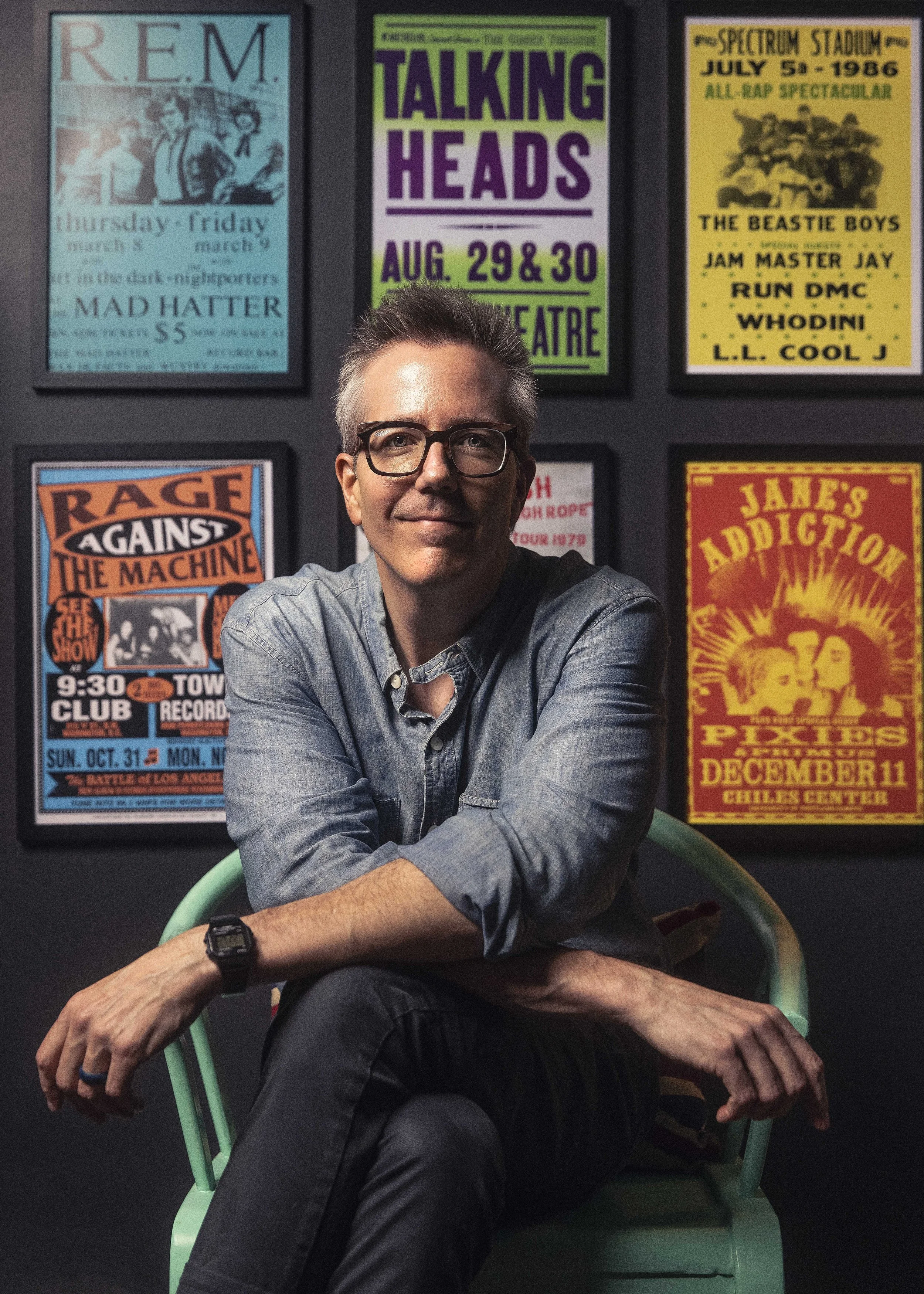 A man with glasses, wearing a denim shirt and a watch, sitting cross-legged on a green chair in front of a wall with colorful concert posters.