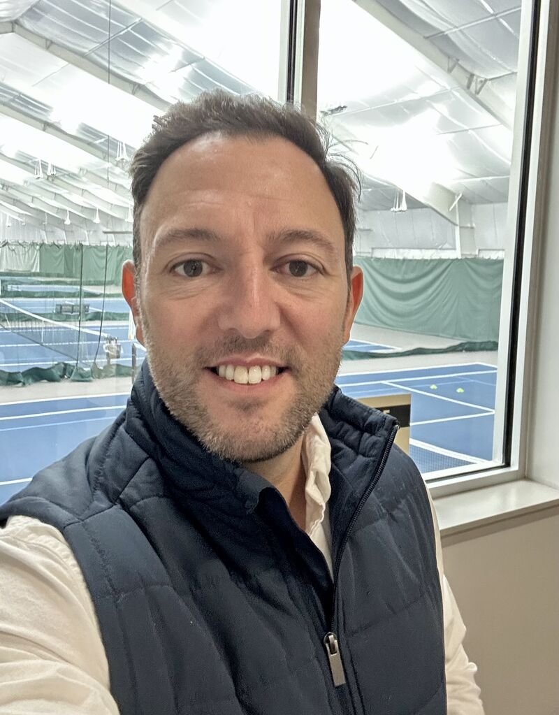 A man taking a selfie near an indoor tennis court with green curtains and tennis balls in the background.