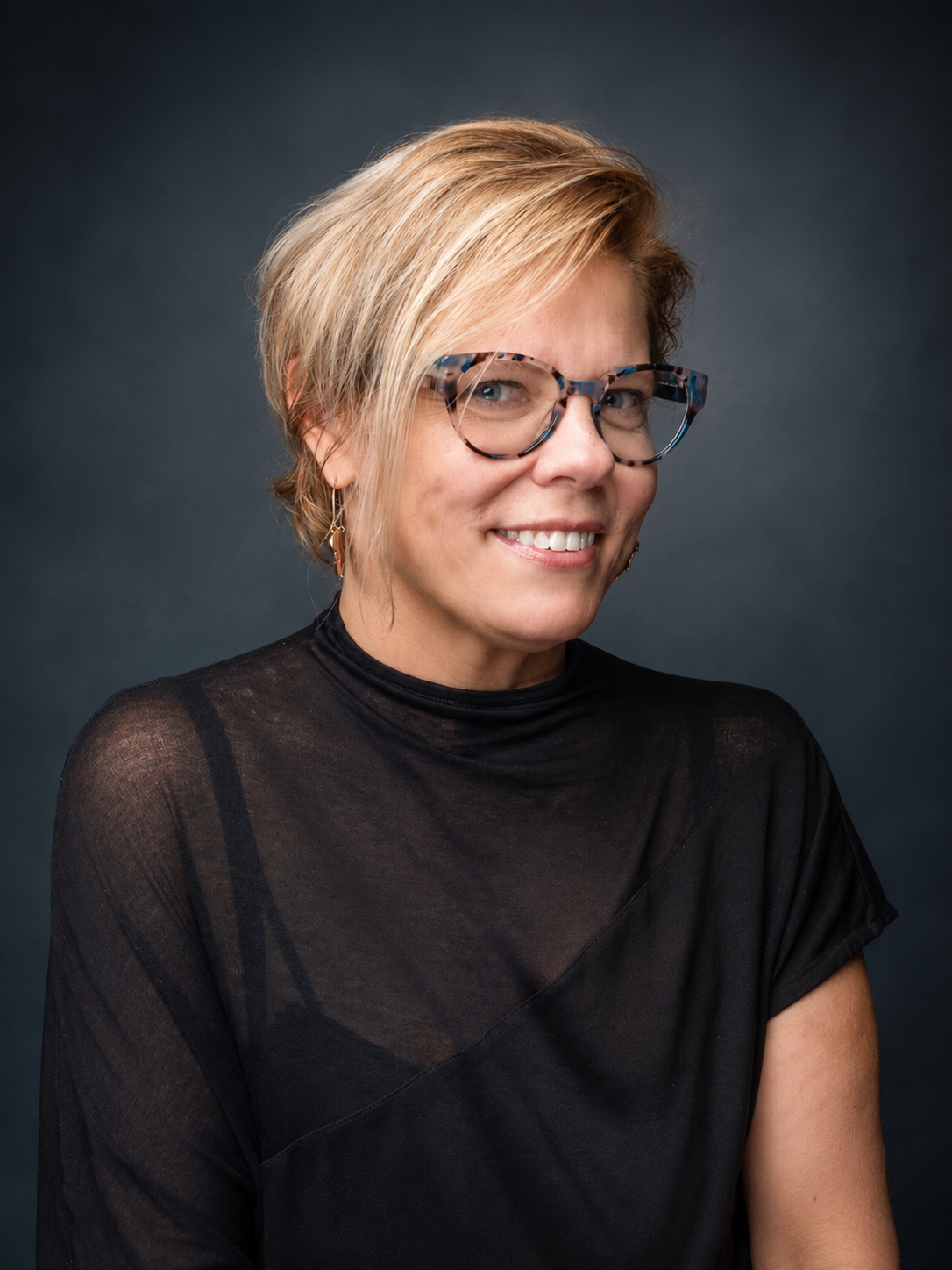 Portrait of a smiling woman wearing glasses and a black top with a dark background.