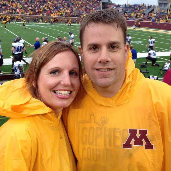 A smiling couple in yellow rain jackets at a football game, with players and spectators in the background on the field.