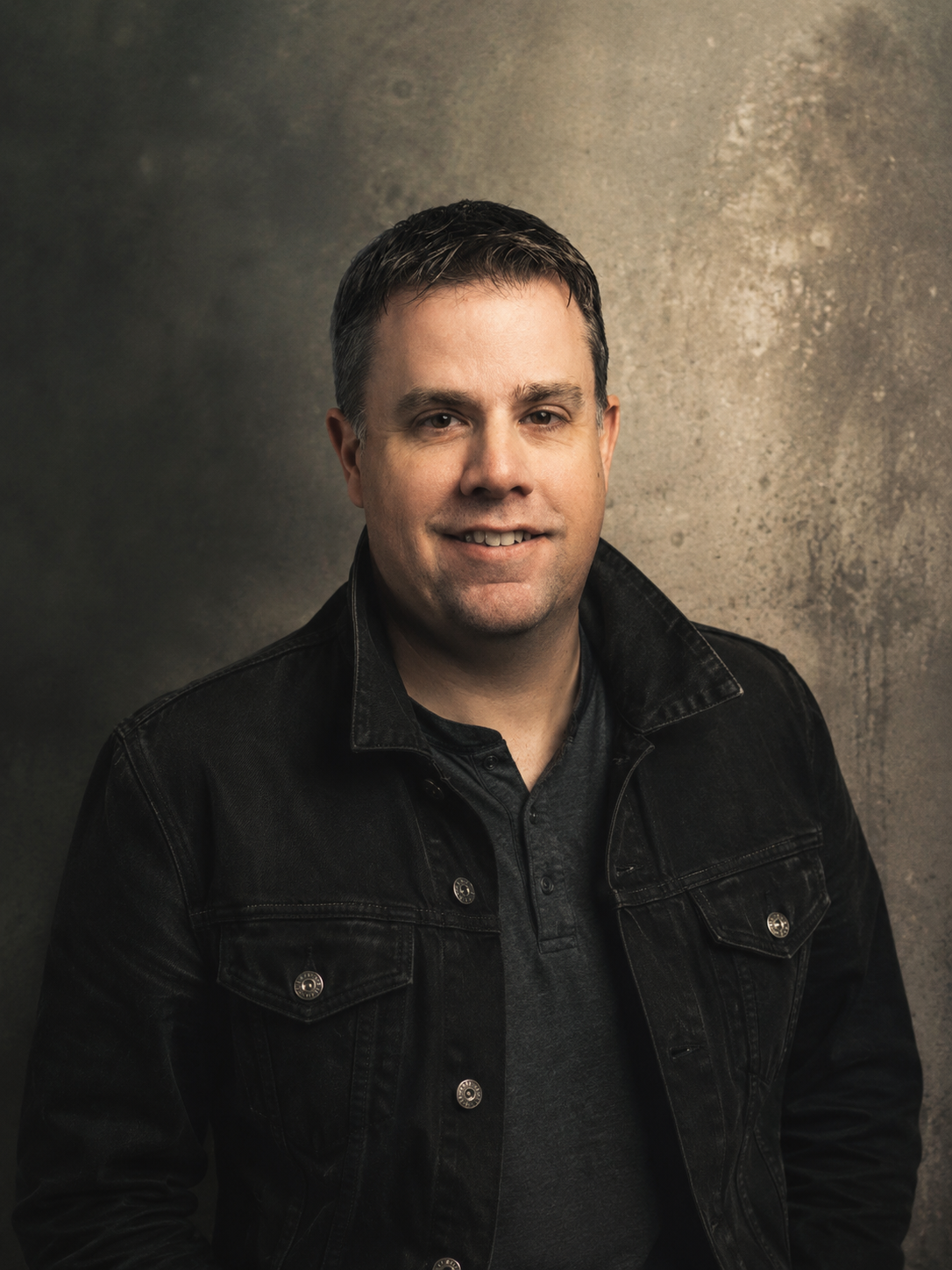 A man with short brown hair, wearing a black denim jacket over a dark shirt, smiling at the camera against a textured gray background.