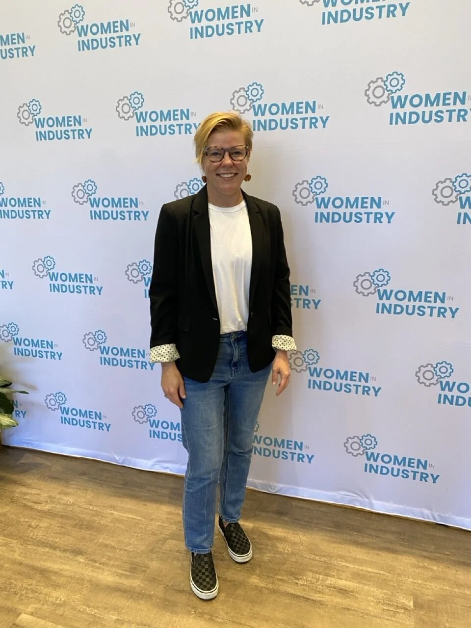 A woman standing in front of a backdrop with the logo and text 'Women in Industry' repeated multiple times. She is smiling, wearing glasses, a black blazer with polka dot cuffs, a white shirt, blue jeans, and checkered slip-on shoes.