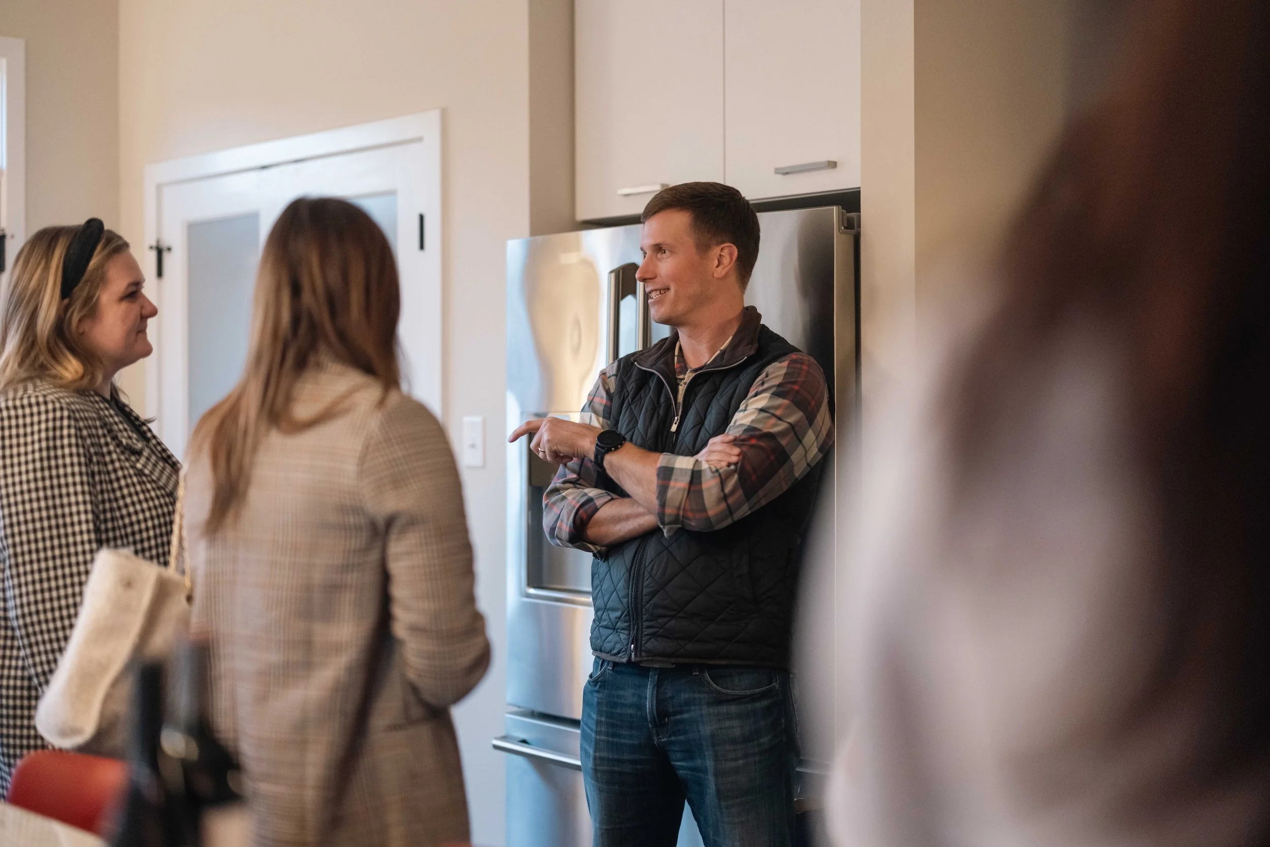 Three women and one man engaged in a conversation in a kitchen. The man is smiling, wearing a black vest and plaid shirt with his arms crossed, and the women are facing him, listening. The kitchen features white cabinets and a stainless steel refrigerator.