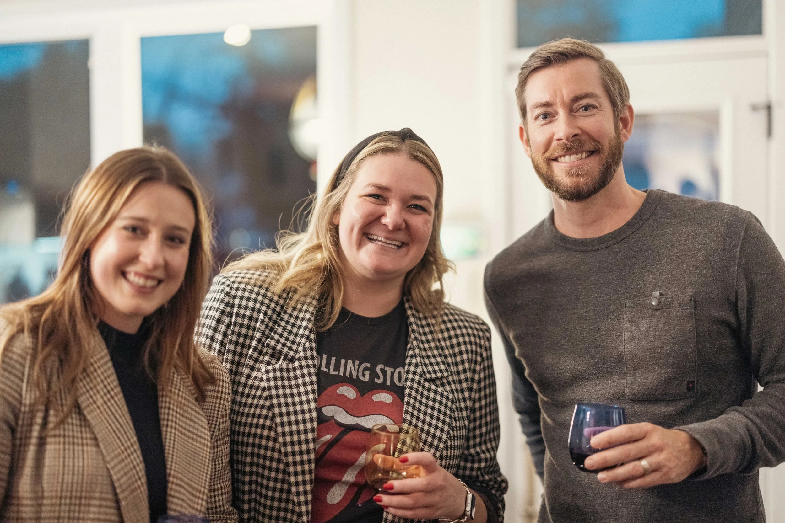 Three people at a social gathering, two women and one man, smiling and holding drinks.