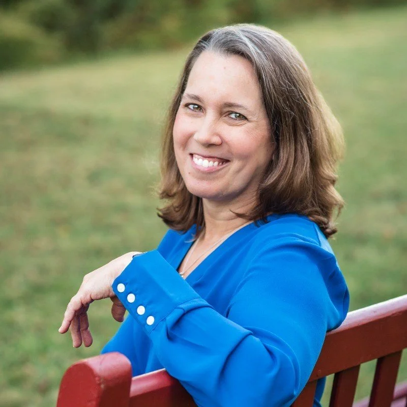 A woman with shoulder-length brown hair, wearing a blue shirt, sitting on a red bench outdoors with a blurred green background, smiling at the camera.