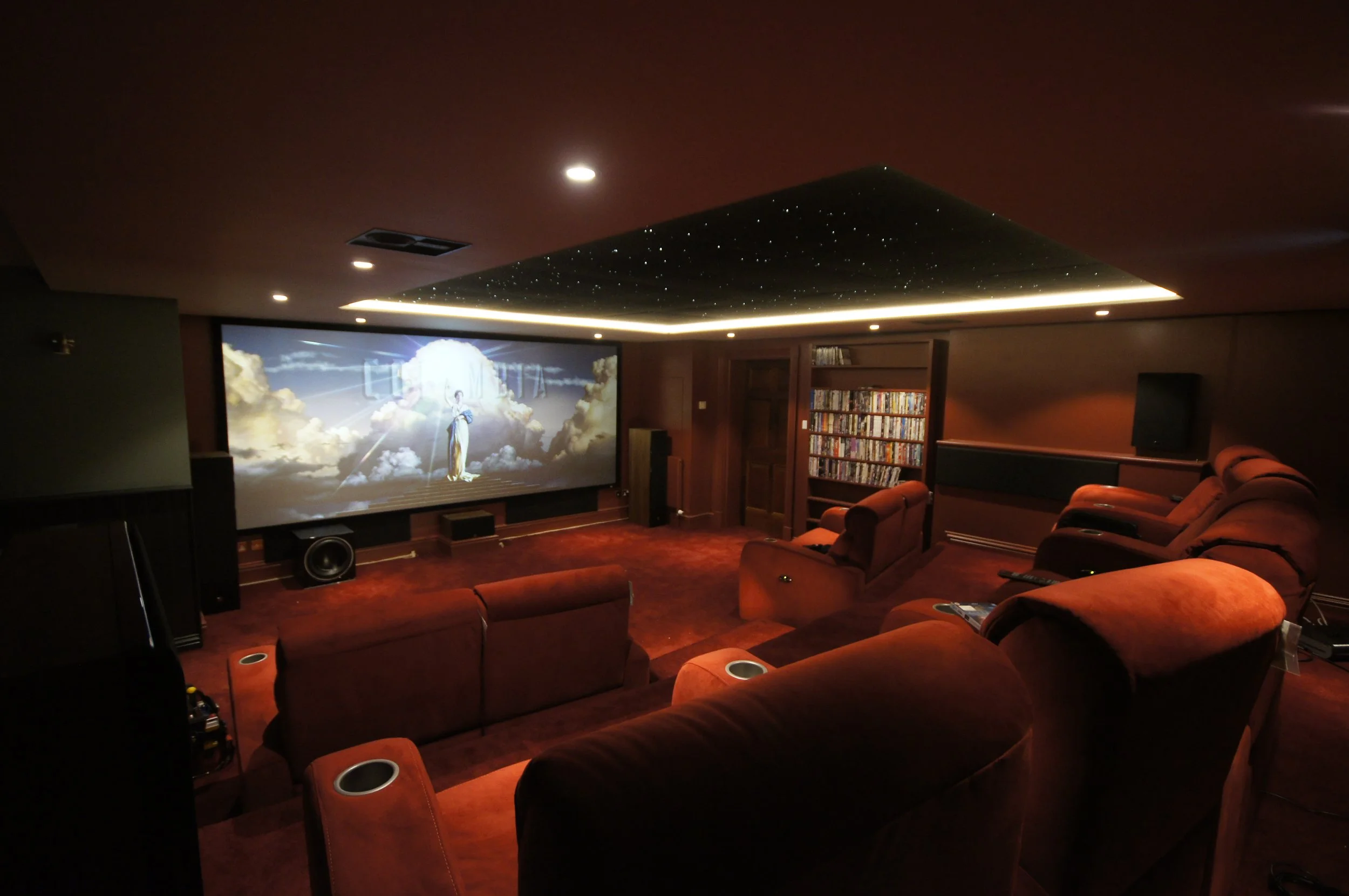A cozy home theater room with multiple red recliner seats, a large screen displaying scenes from a movie, wooden shelves filled with DVDs, and dark ceiling with star-like light fixtures.