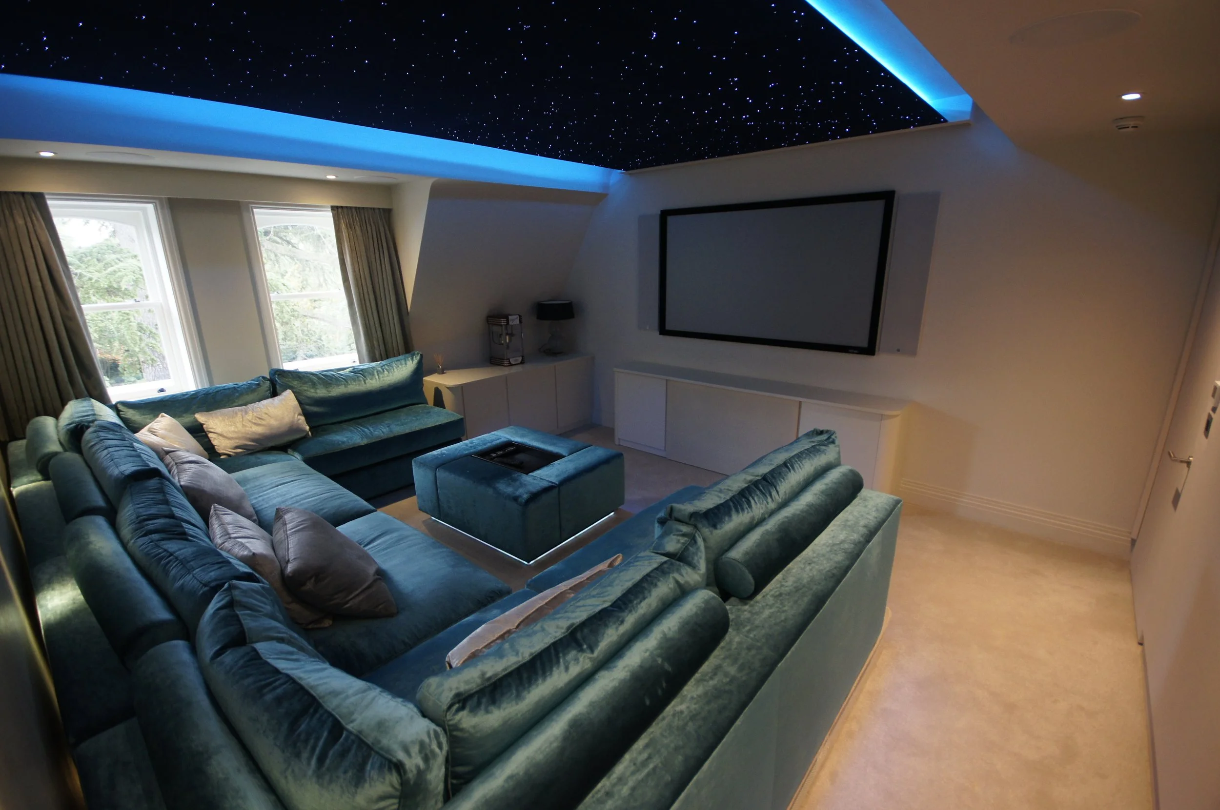 Modern living room with a large, plush teal sectional sofa, multiple throw pillows, and a ceiling decorated with glowing star-like lights and blue LED accents. Large windows let in natural light and reveal a view of trees outside.
