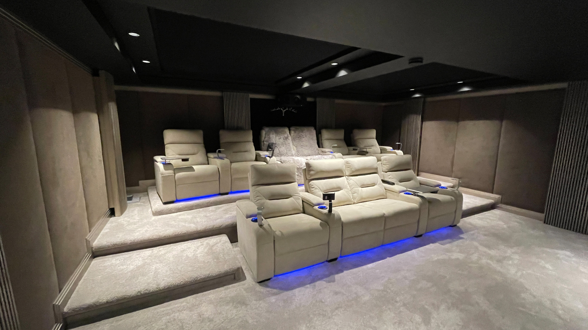 Luxury home theater room with cream-colored leather recliner seats arranged on steps, featuring blue LED lighting underneath, dark walls, and a black ceiling with recessed lighting.