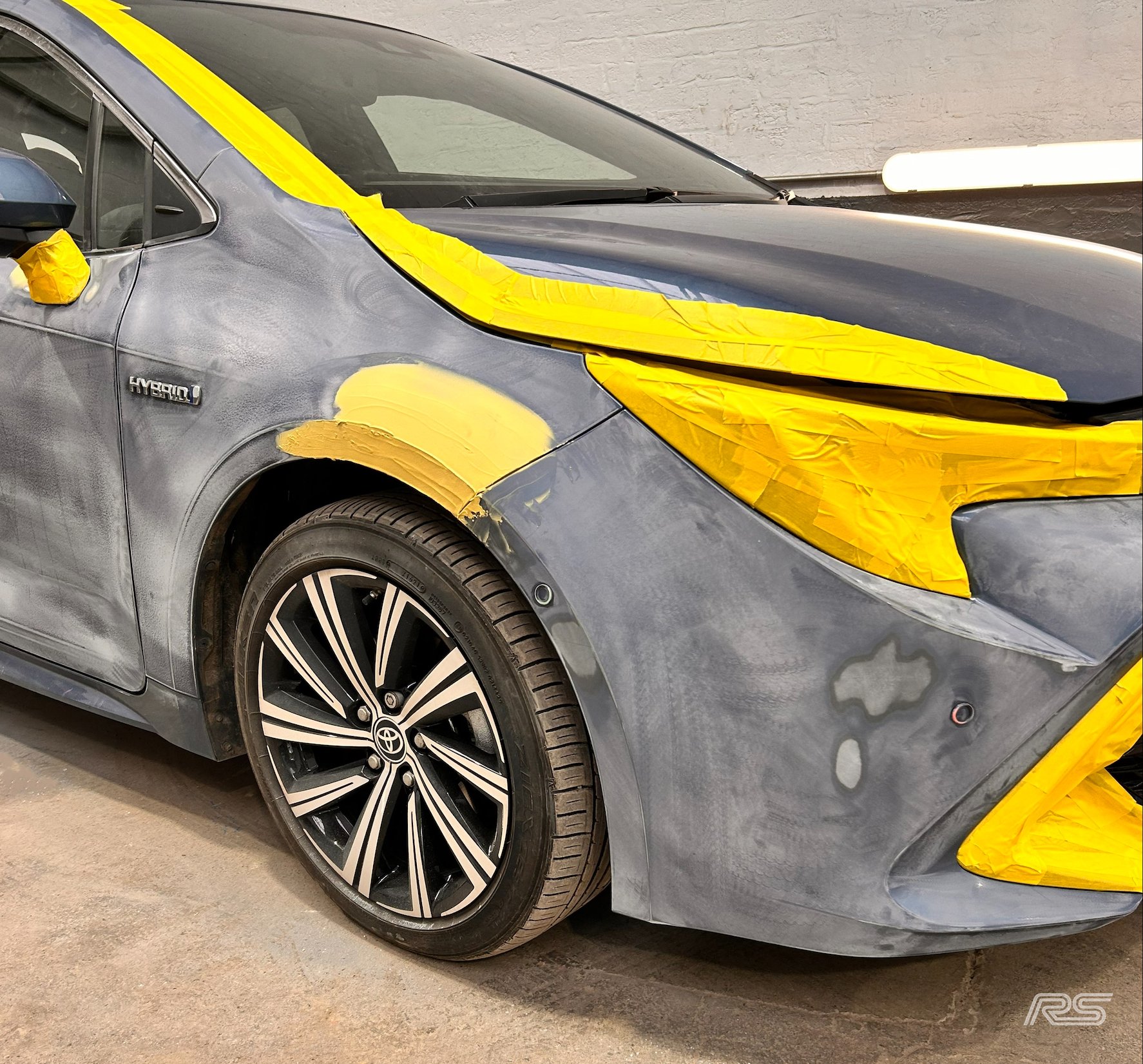A Toyota car in a workshop with yellow painter's tape covering the windshield, headlights, and other parts in preparation for painting & repair.