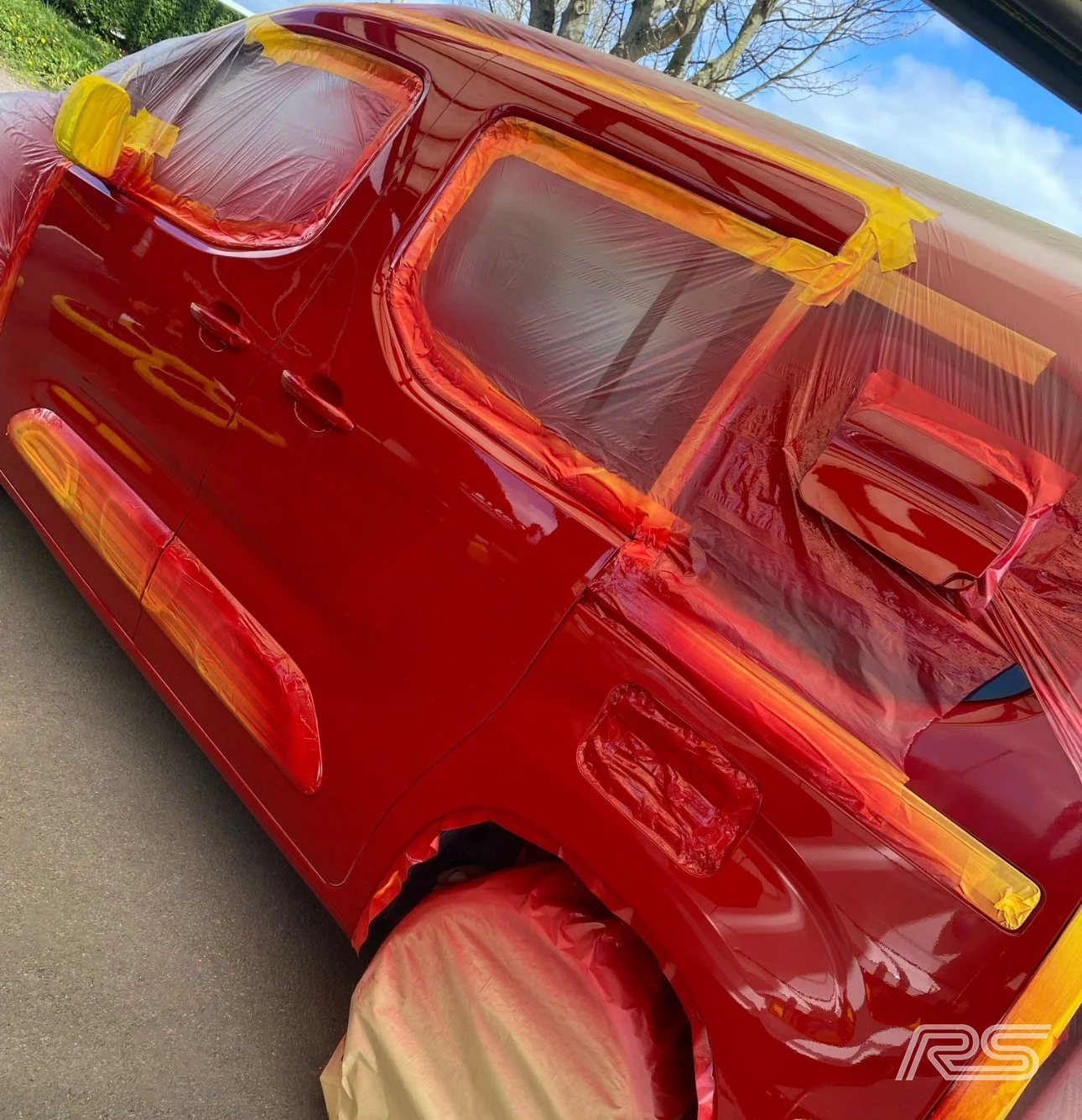 A red car covered in masking tape and plastic sheeting for painting, with one front wheel covered with masking paper, parked outdoors near some trees.