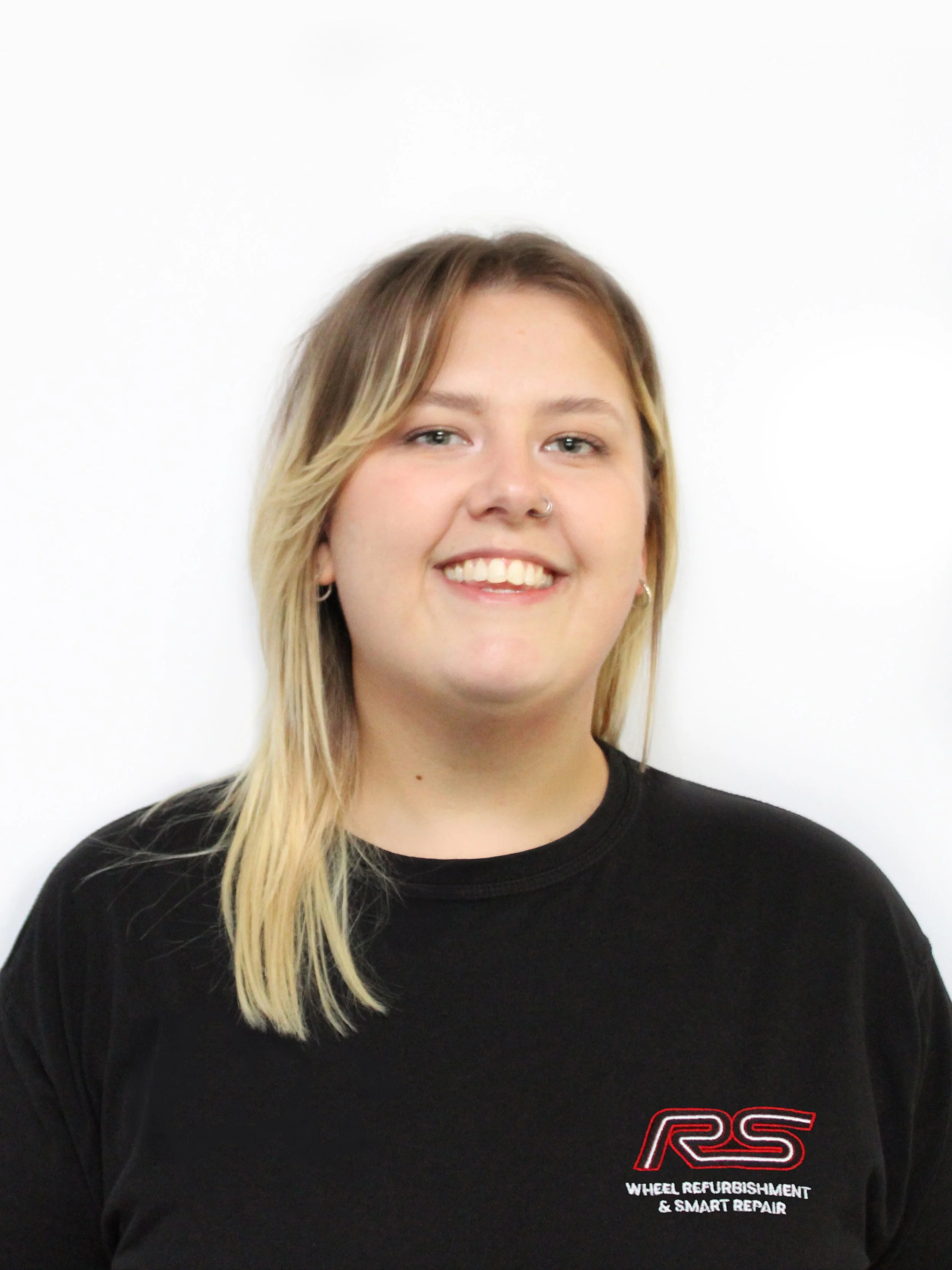 Portrait of a young woman with shoulder-length blonde hair, smiling, wearing a black t-shirt with a logo that reads 'RS Wheel Refurbishment & Smart Repair,' standing against a white background.