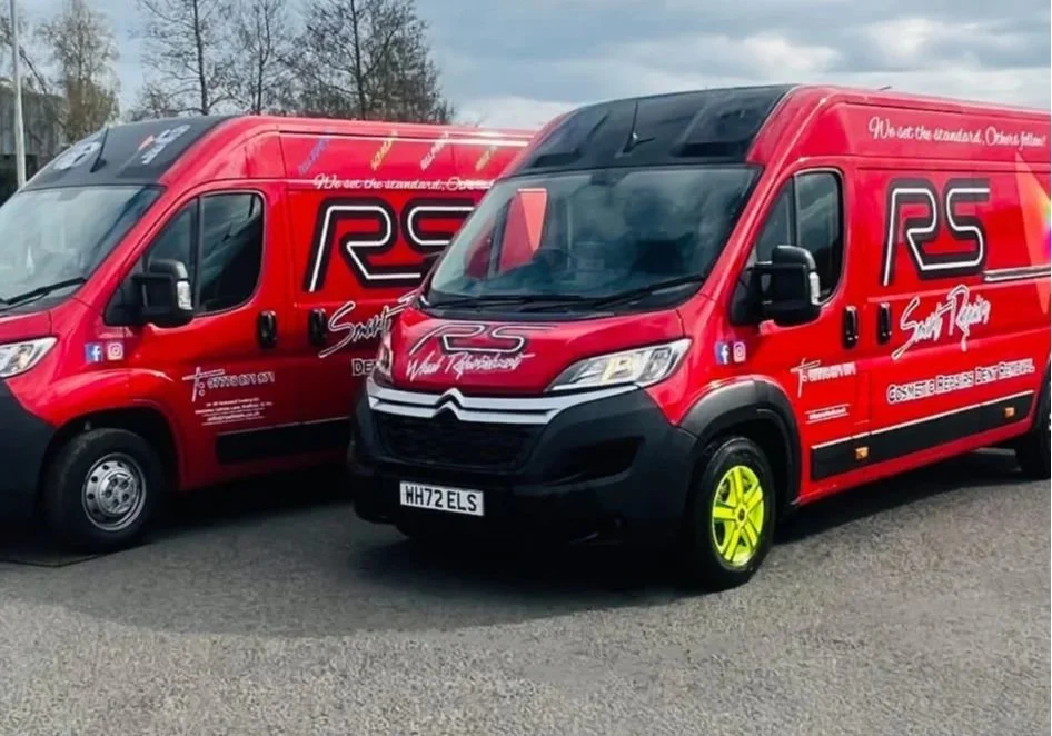 Two red vans with branding and logos for RS Wheels, parked side by side on a paved surface, with one van’s front wheel painted bright yellow and various text and social media icons on the sides.