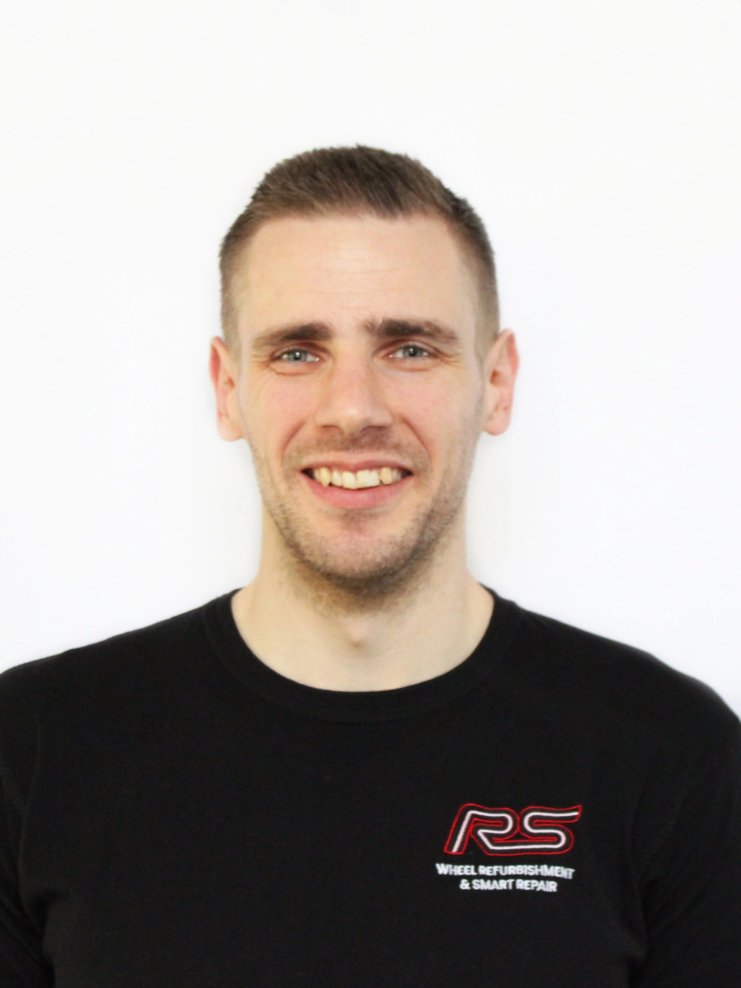 A smiling man with short brown hair and a black t-shirt that has a RS logo and the words 'Wheel Refurbishment & Smart Repair'.