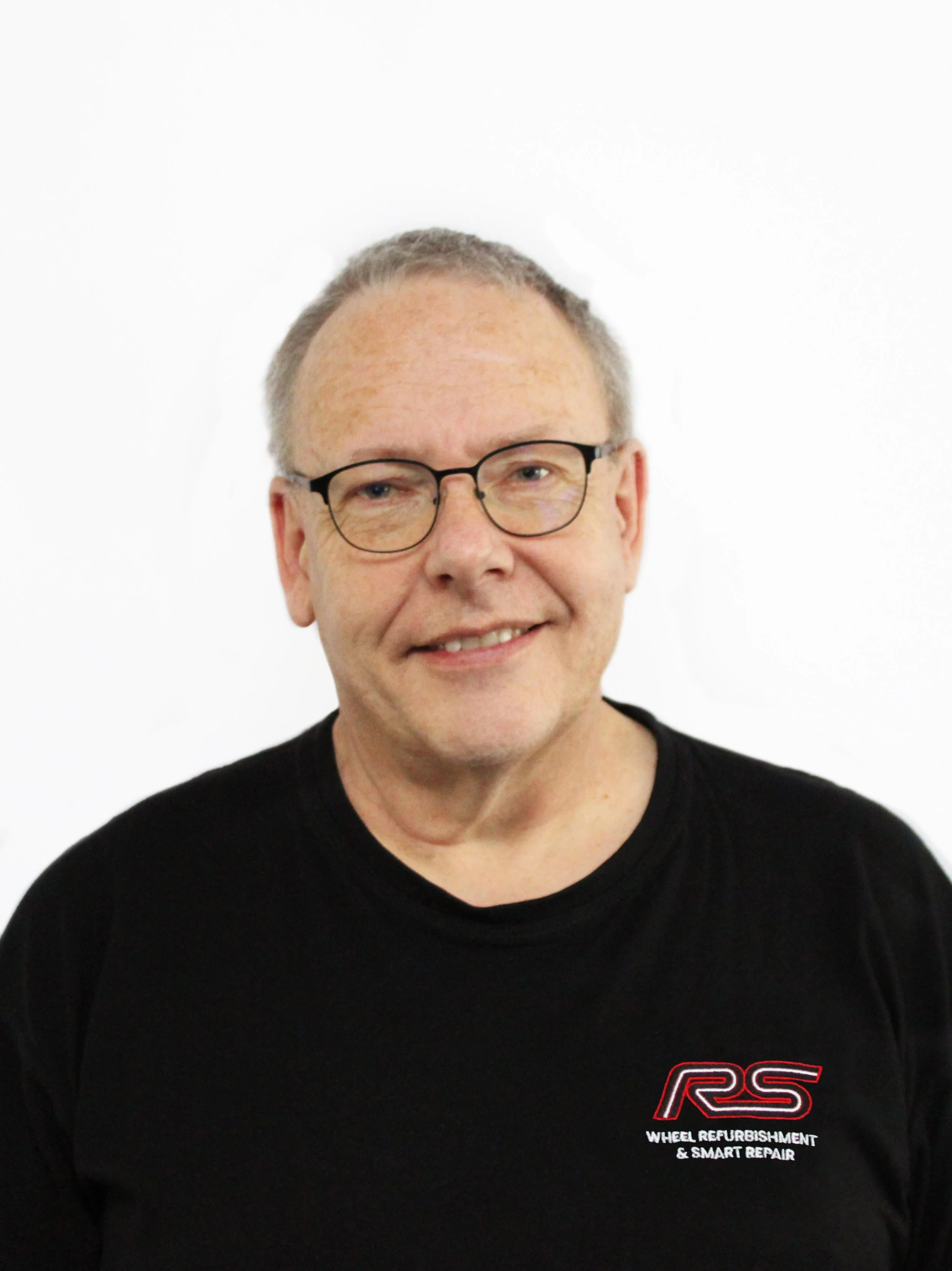 Portrait of a middle-aged man with glasses, wearing a black t-shirt with a logo and text that reads 'Wheel Refurbishment & Smart Repair'.