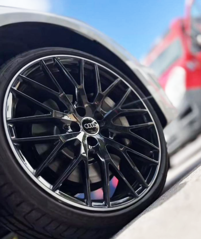 Close-up of a black Audi alloy wheel with the Audi logo at the center. The wheel is mounted on a car, with a blue sky background and part of a red and gray vehicle visible.