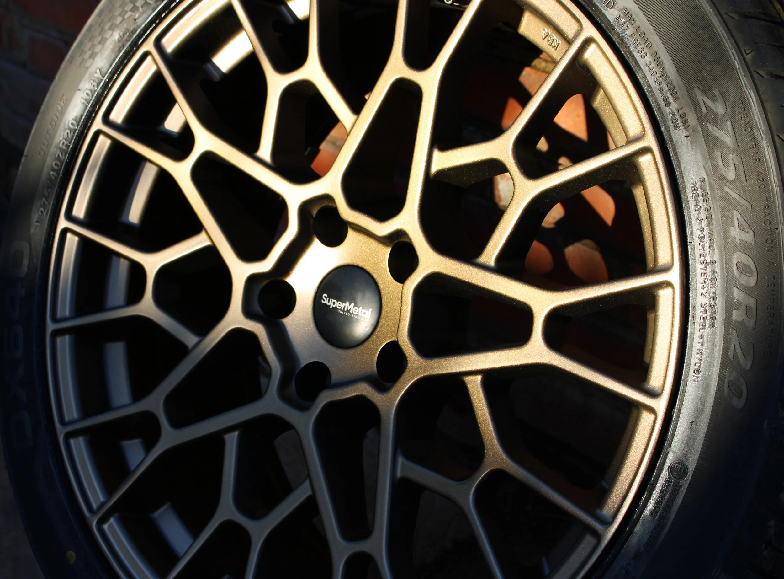Close-up of a multi-spoke alloy in a performance bronze with a satin finish. The center of the rim has a black hubcap with white text reading "SuperMetal."