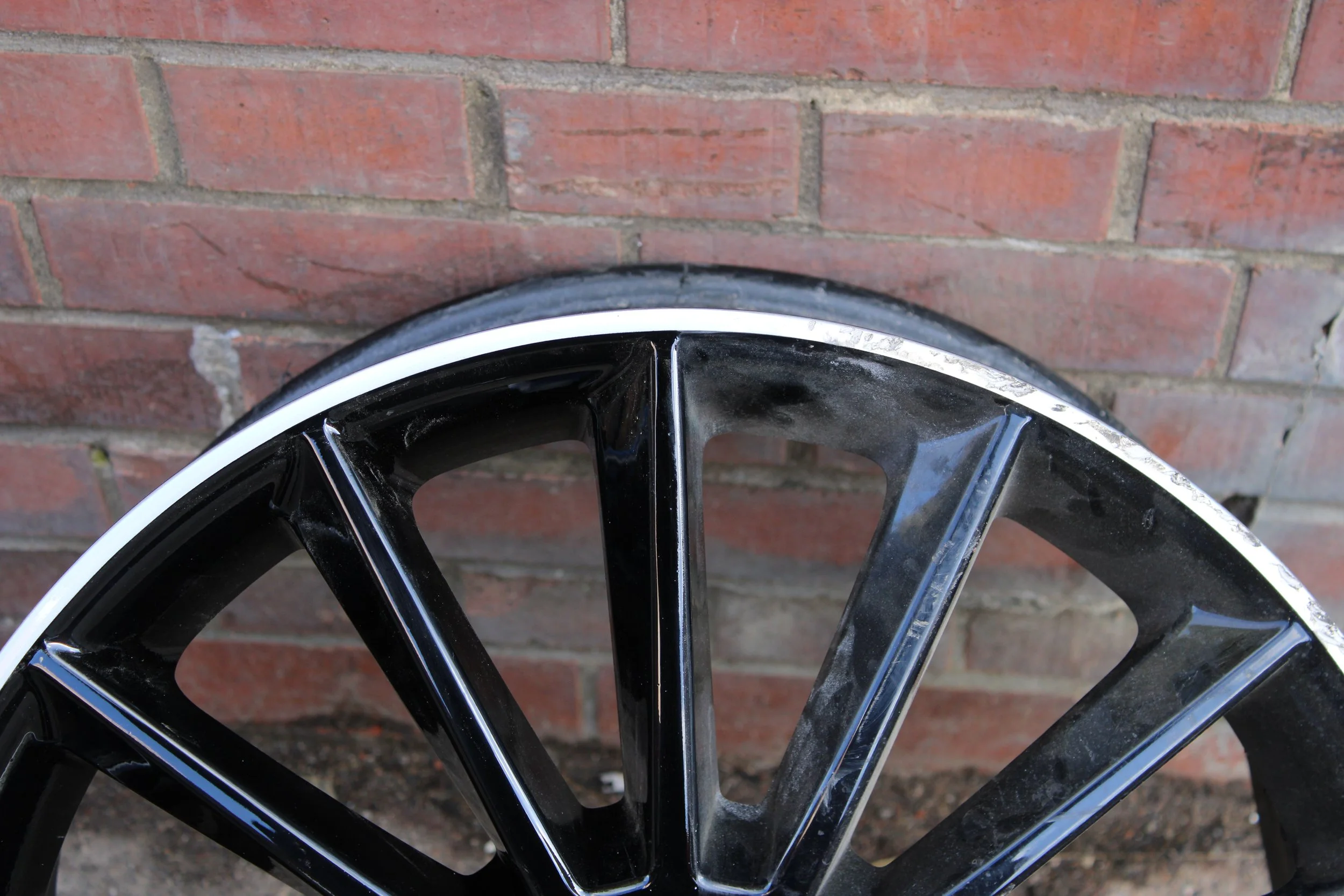 Close-up of a black alloy car wheel with a silver rim, leaning against a red brick wall. the left half of the wheel has been repaired and is new and shiny while the right is still scuffed and damaged.