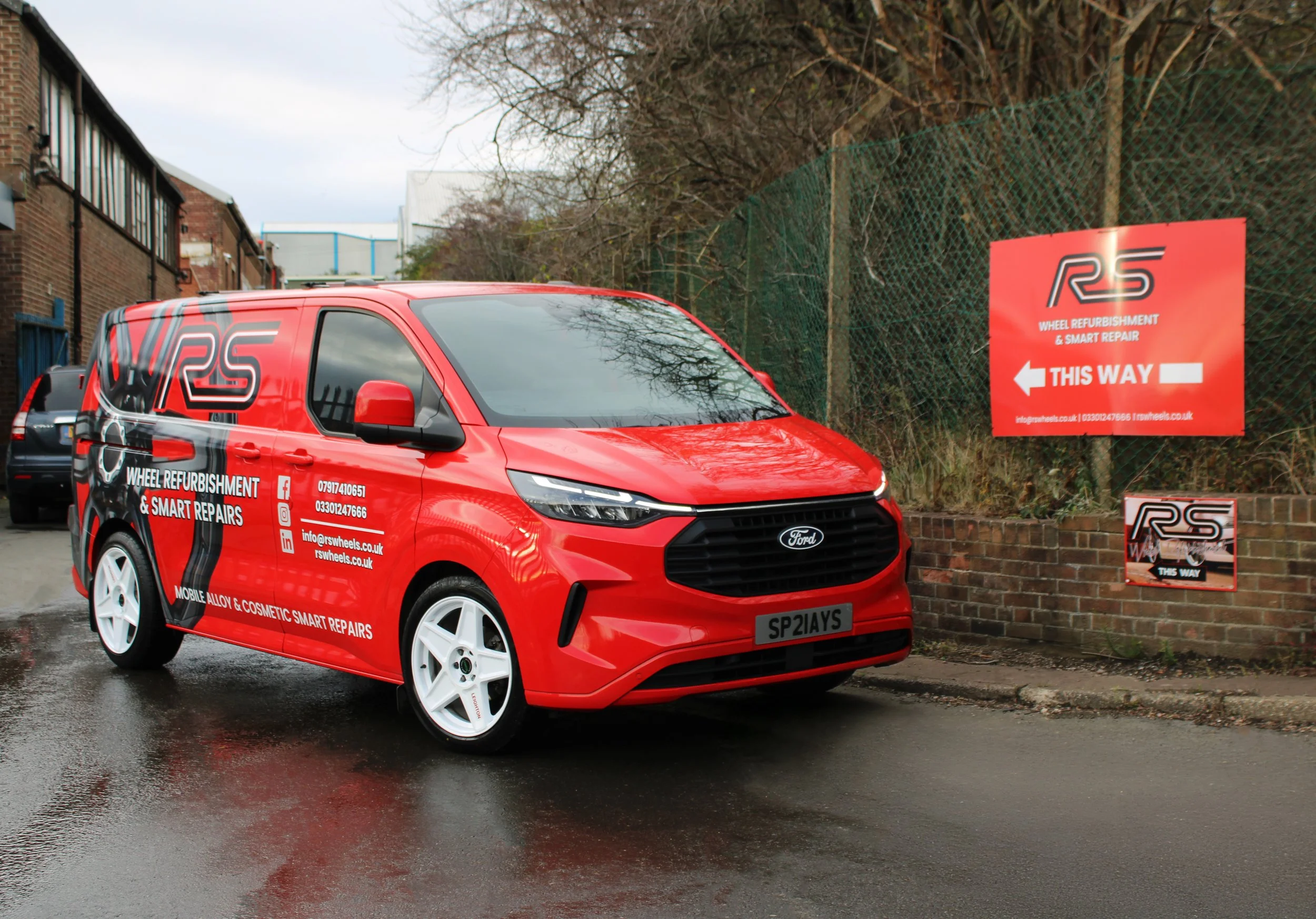 Red van advertising wheel refurbishment and smart repairs parked on a street near a red sign pointing the way to RS Wheel Refurbishment. The van has white wheels and various contact details printed on the side.