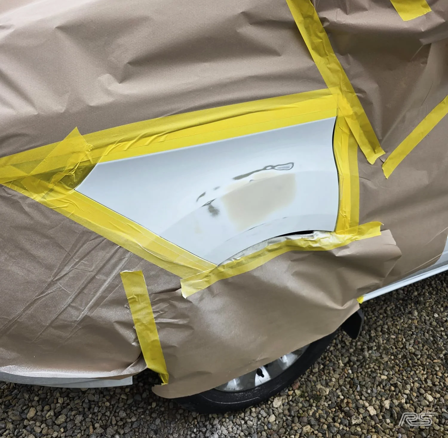 A car with a section wrapped in brown paper and yellow painter's tape, revealing a small window on the side that is being repaired.