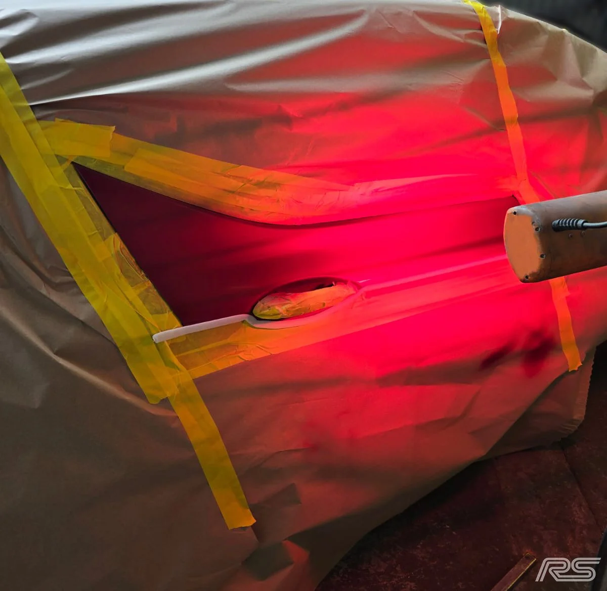 A car undergoing a paint curing process with heat lamps shining on the vehicle, which is covered with protective plastic and taped areas.