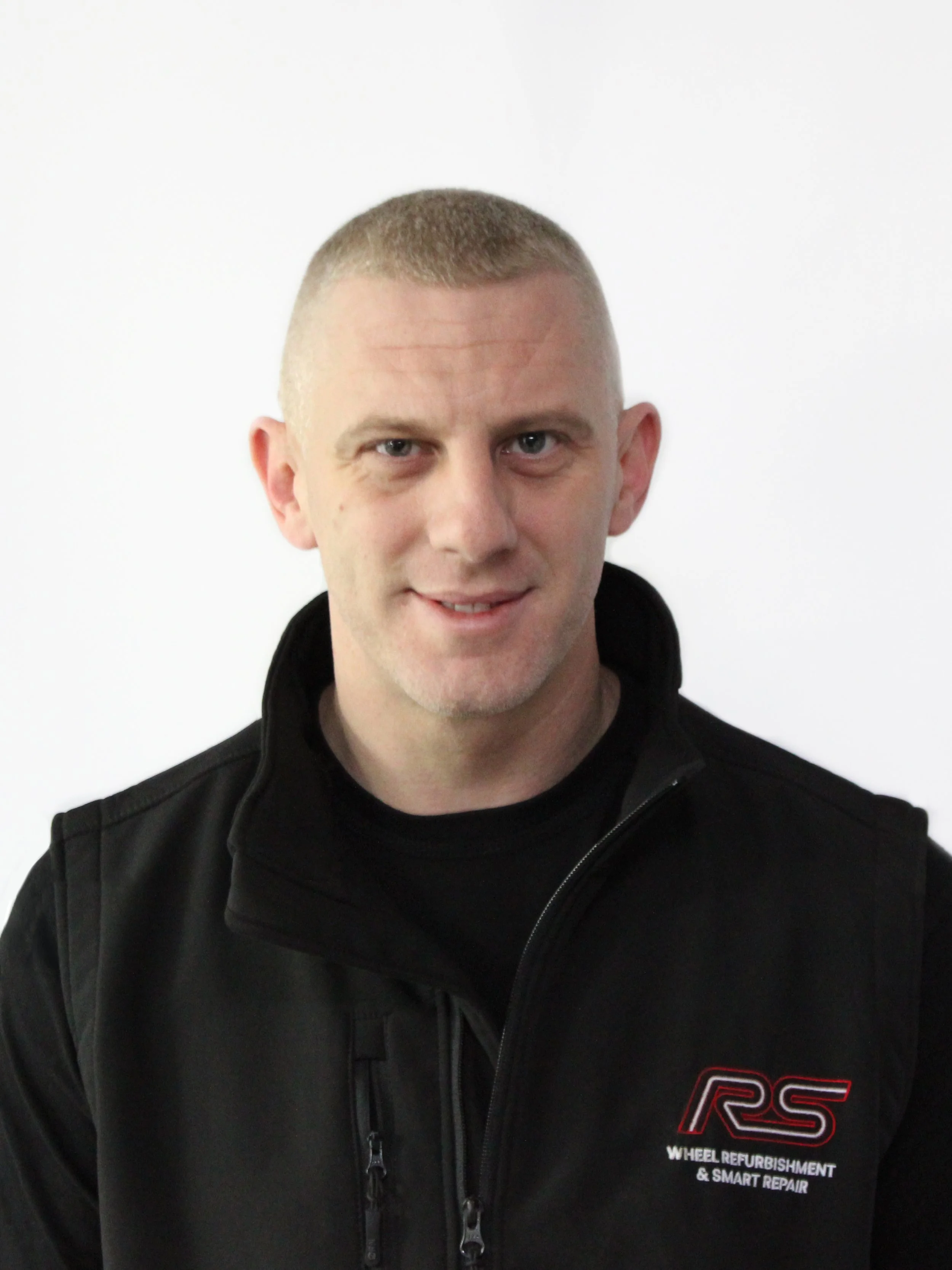 A man with short blonde hair, wearing a black jacket with a logo on it, smiling at the camera against a plain white background.