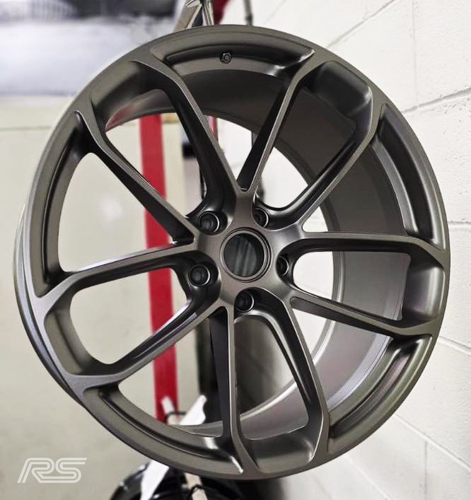 Close-up of a grey alloy with a modern, multi-spoke design, hanging on a rack in the middle of a powder coat refurbishment