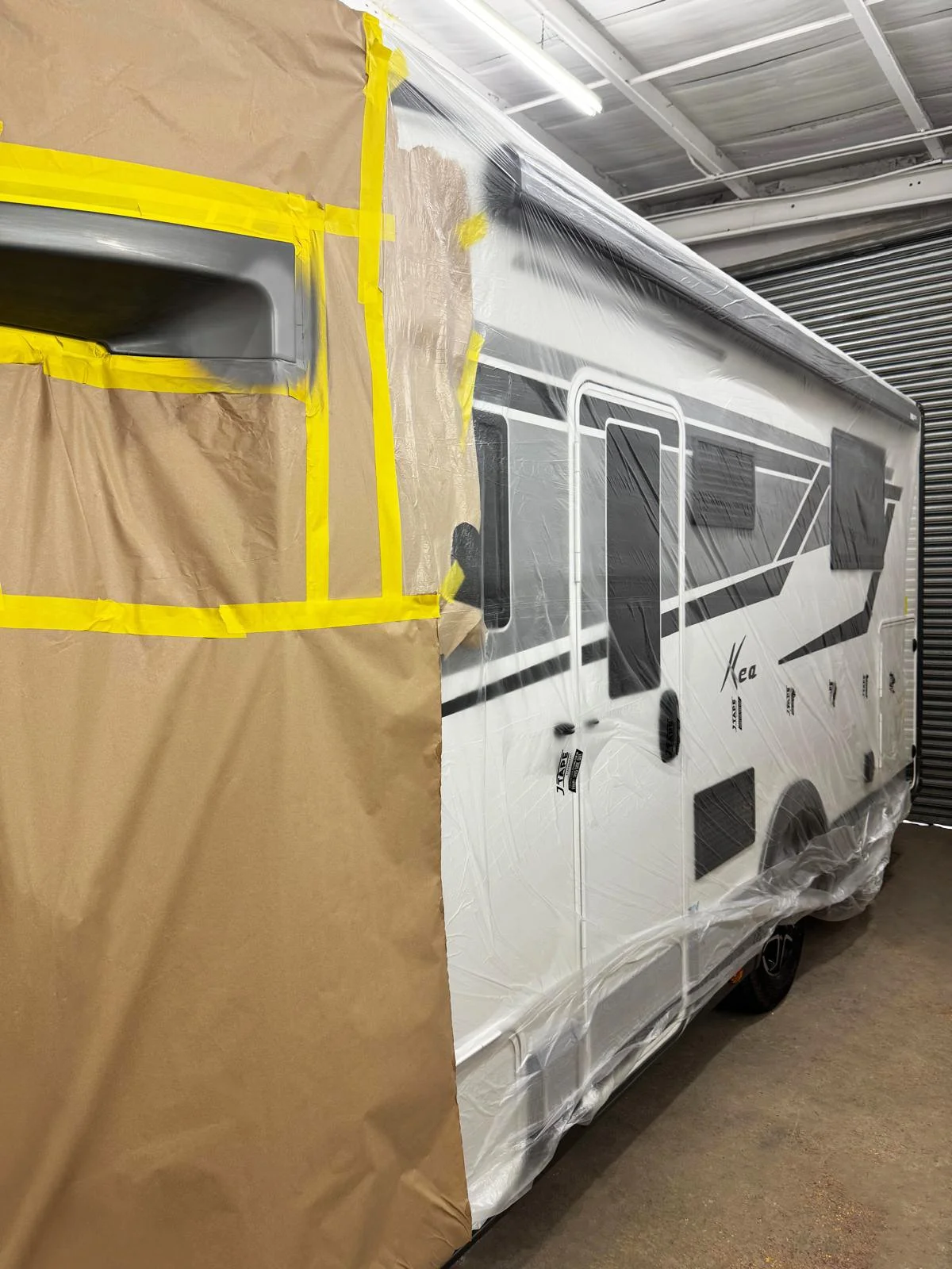 A recreational vehicle (RV) inside a garage, partially covered with plastic sheeting and masking tape for repair and painting.